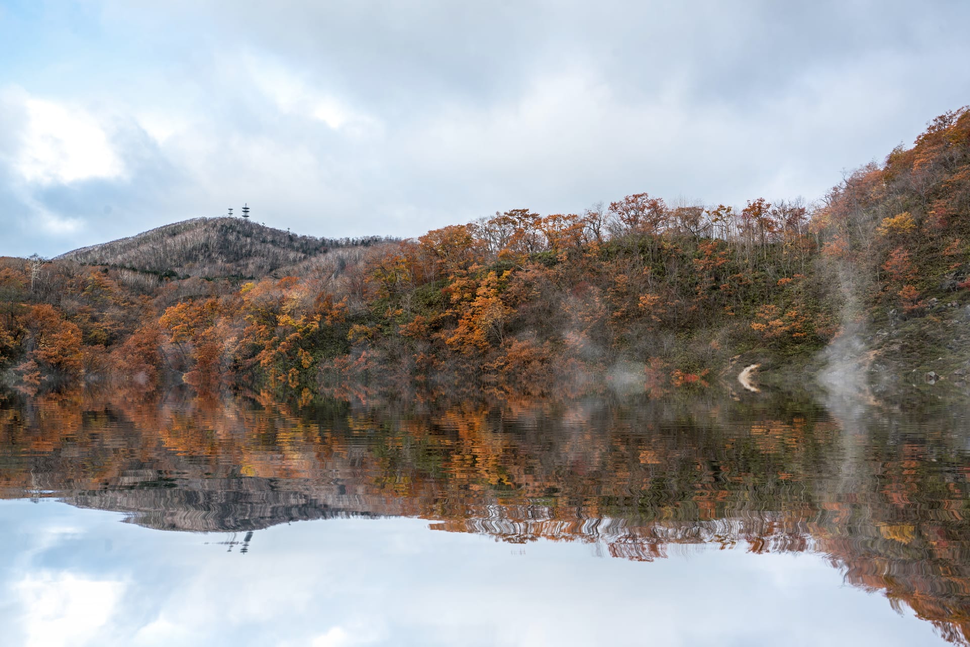 Autumn hillsides reflected in still water, steam rising from a hot spring