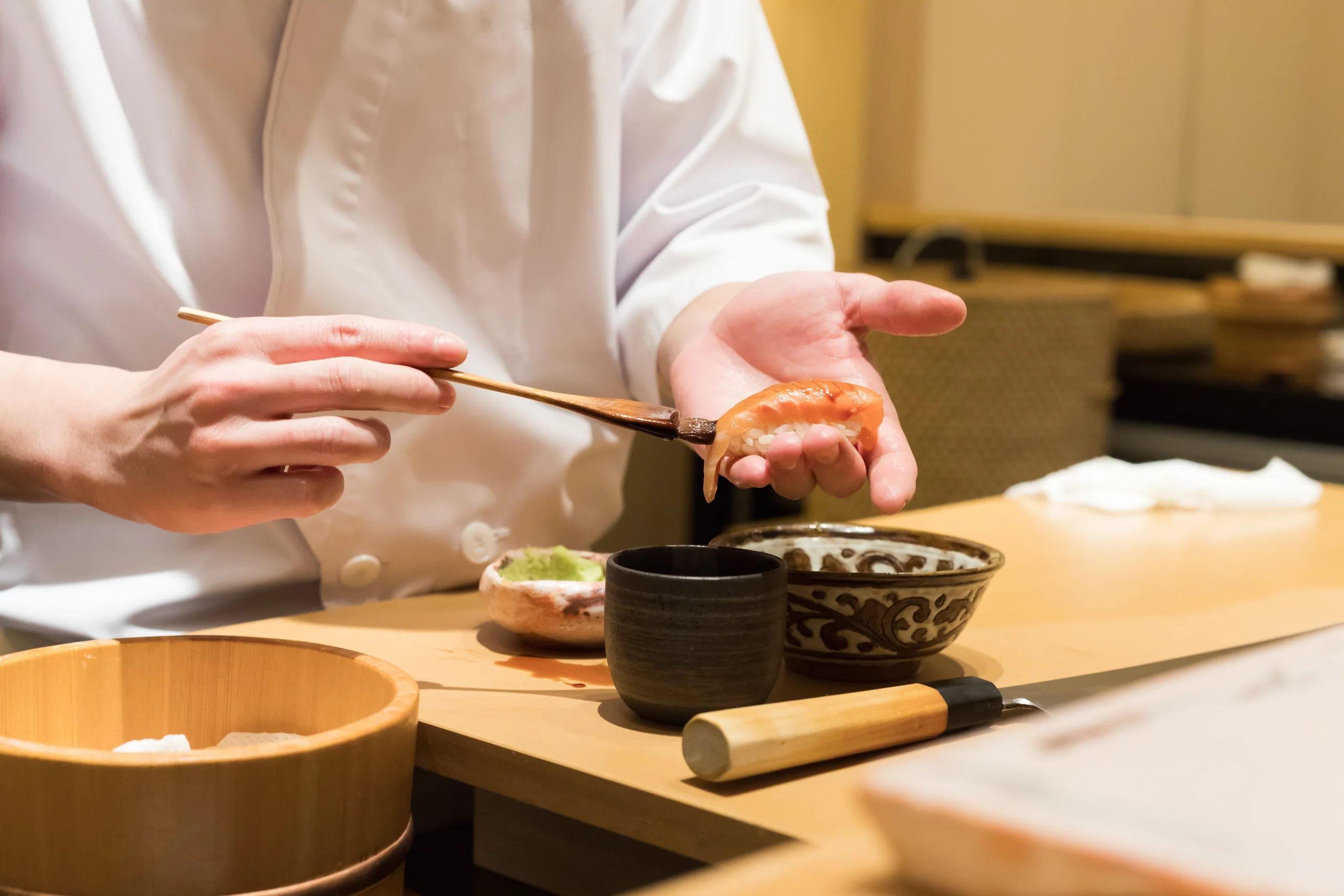 A sushi chef presenting a piece of shrimp nigiri in his palm across the counter