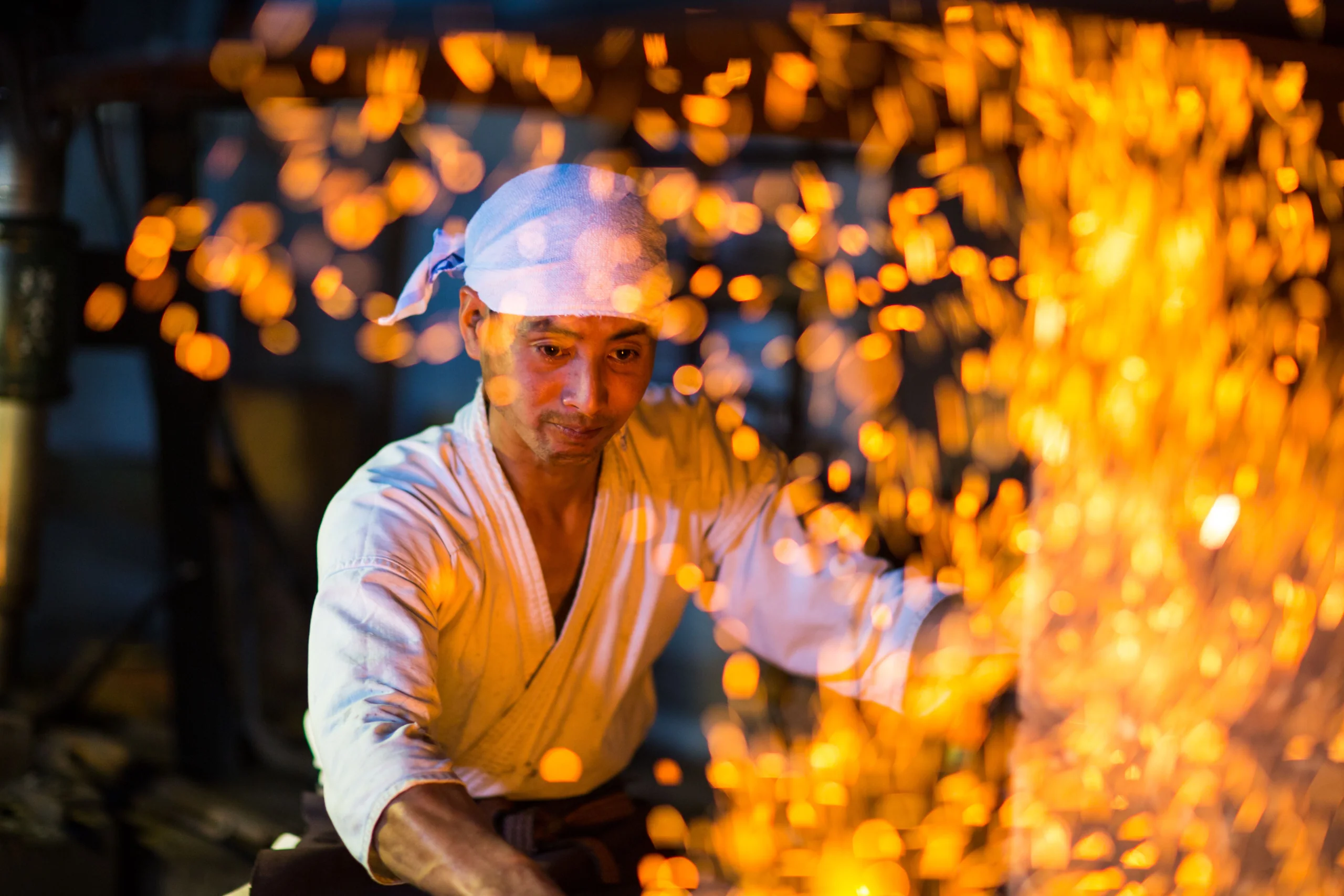 A swordsmith in white work clothes amid a shower of sparks from the forge