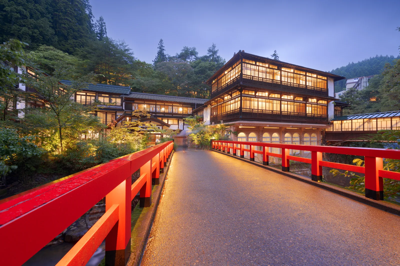 A wooden ryokan lit at dusk beside a red-railed bridge, wooded hills behind