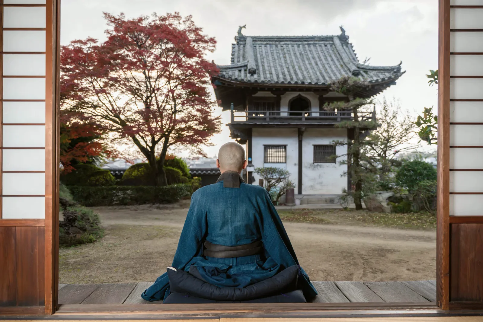 A robed monk seated in meditation on a temple veranda, a maple and a temple hall beyond