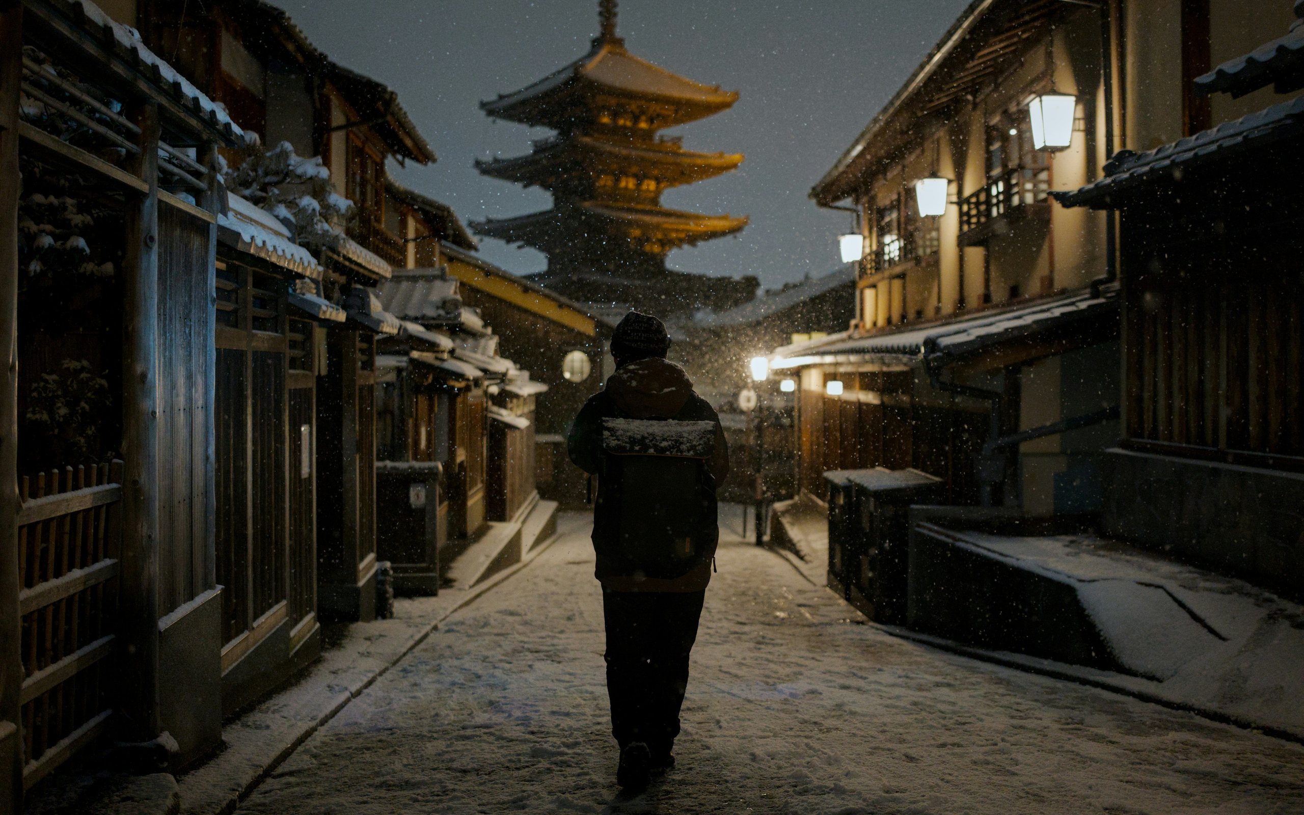 A traveller walking up a snow-dusted lane of wooden houses towards the lit five-storey pagoda of Higashiyama, Kyoto, at night