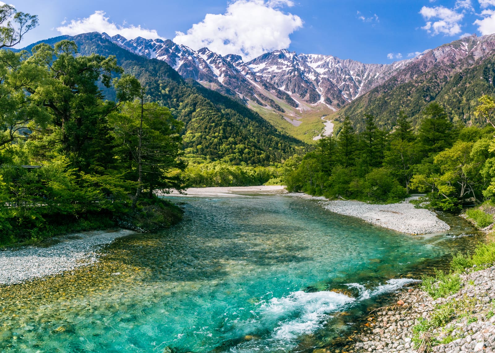 A clear turquoise river running between green forest and snow-streaked peaks of the Japan Alps at Kamikochi
