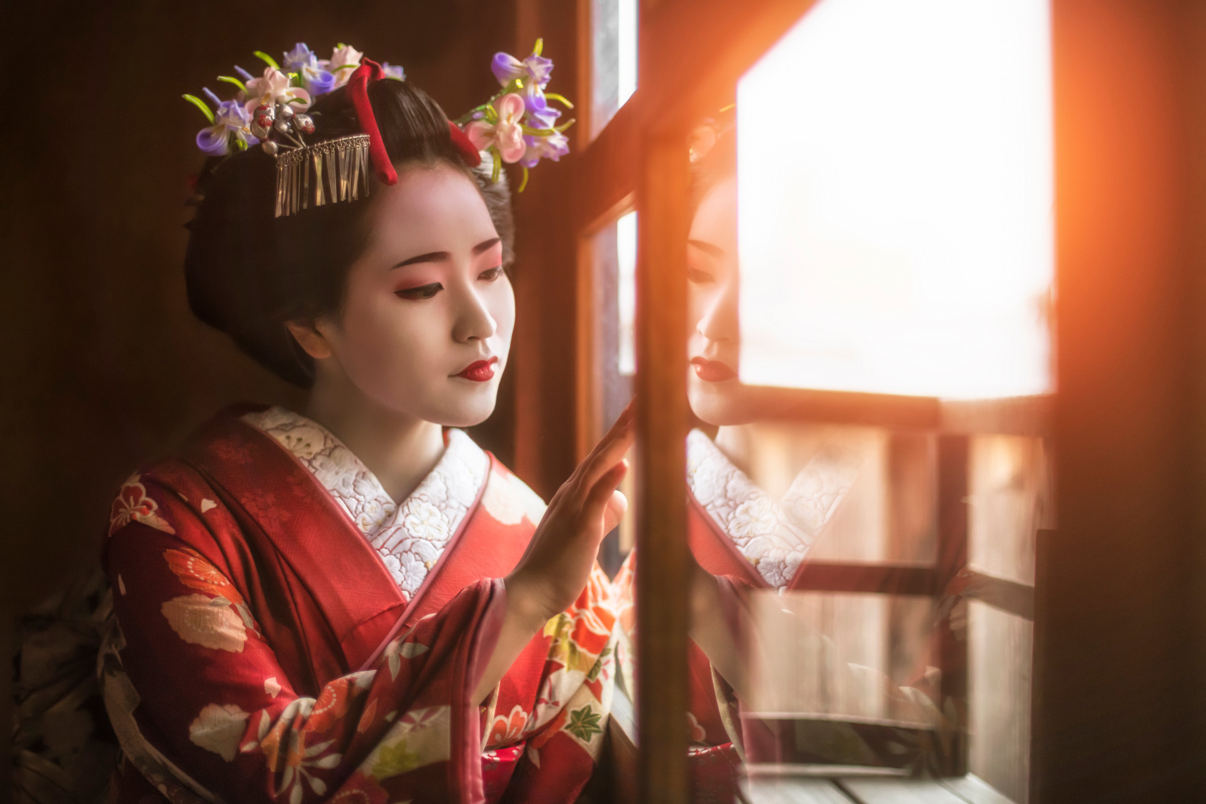 A maiko in a red floral kimono and flowered kanzashi, one hand on a sunlit window pane, her reflection in the glass