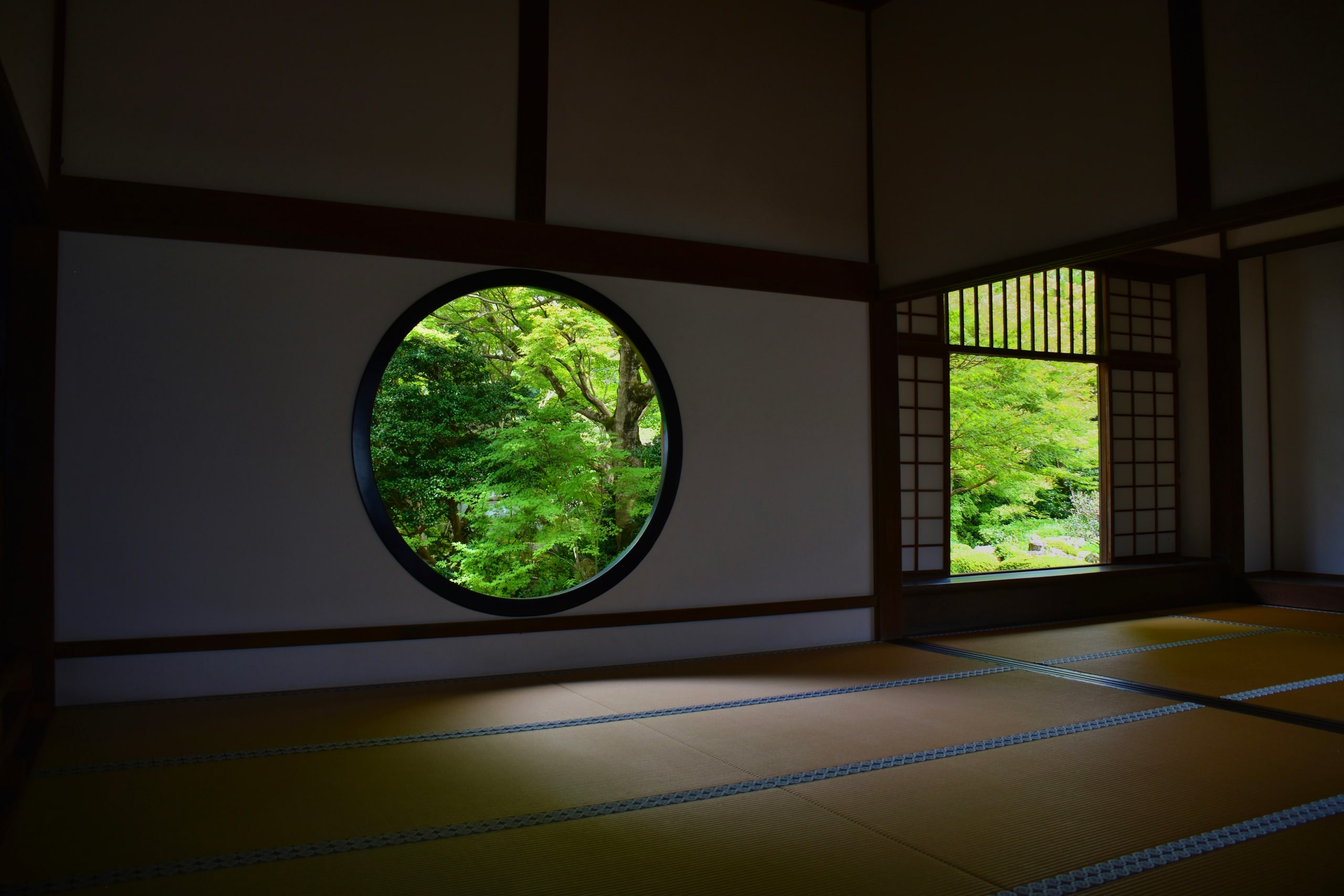A darkened tatami room whose round window opens onto early-summer maples
