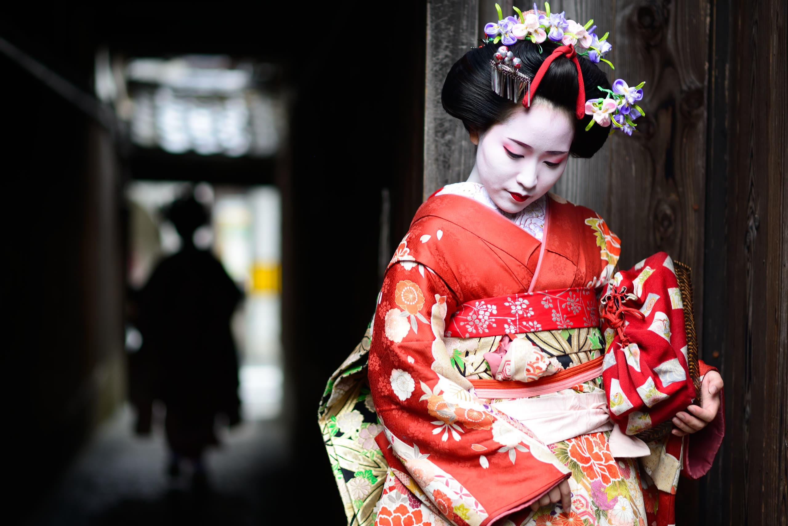 A maiko in a vermillion kimono, eyes lowered, in the dim corridor of a Kyoto machiya