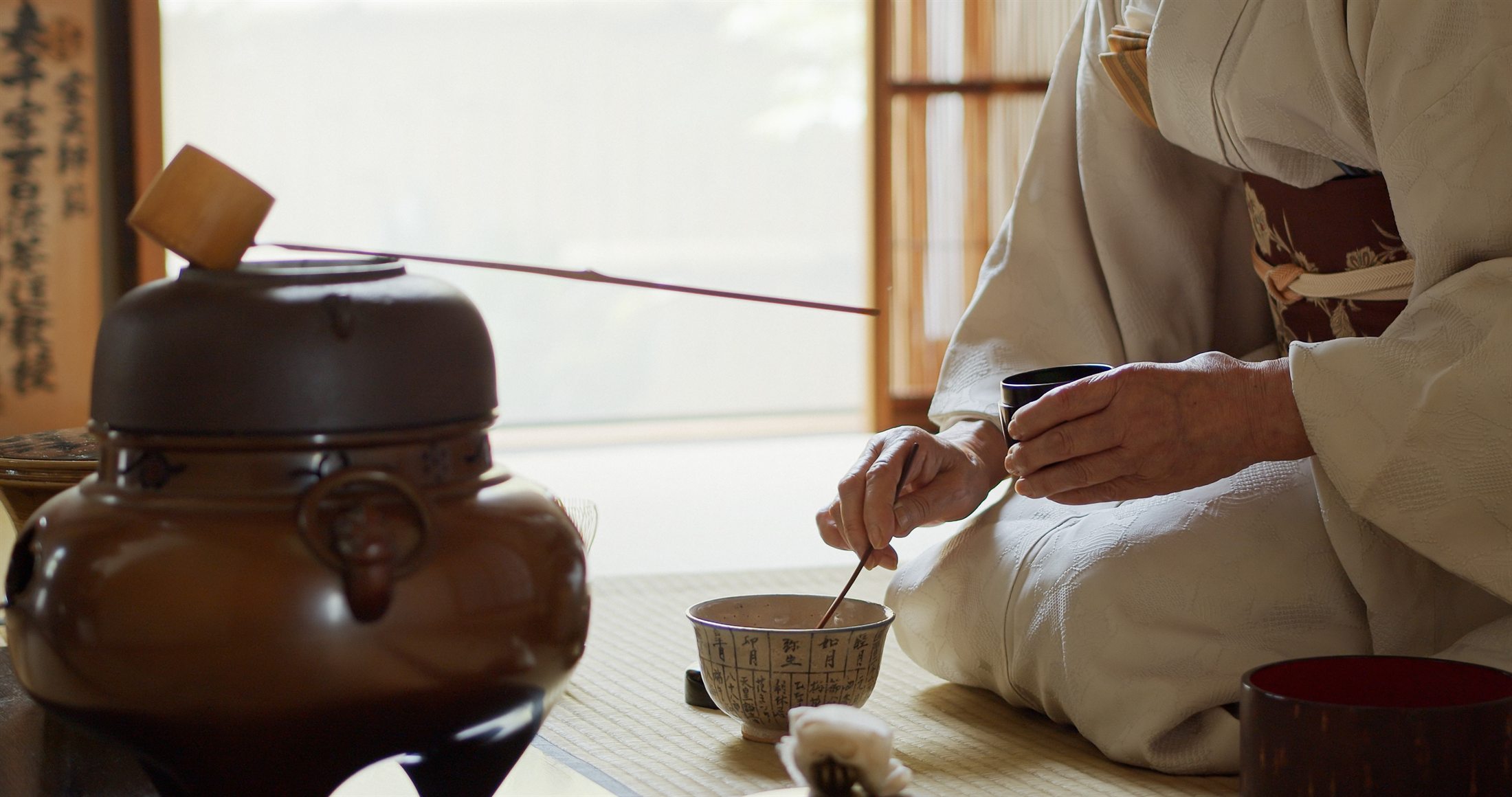A tea ceremony host in a pale kimono, kneeling on tatami, measuring matcha into a bowl beside the kettle