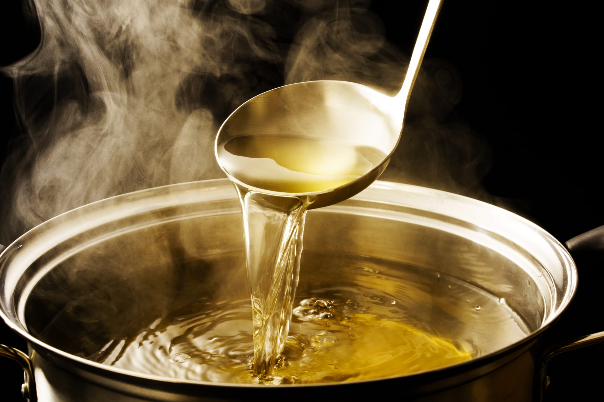 A steel ladle pouring a stream of clear golden dashi back into a steel stockpot, steam rising from the surface against a dark background