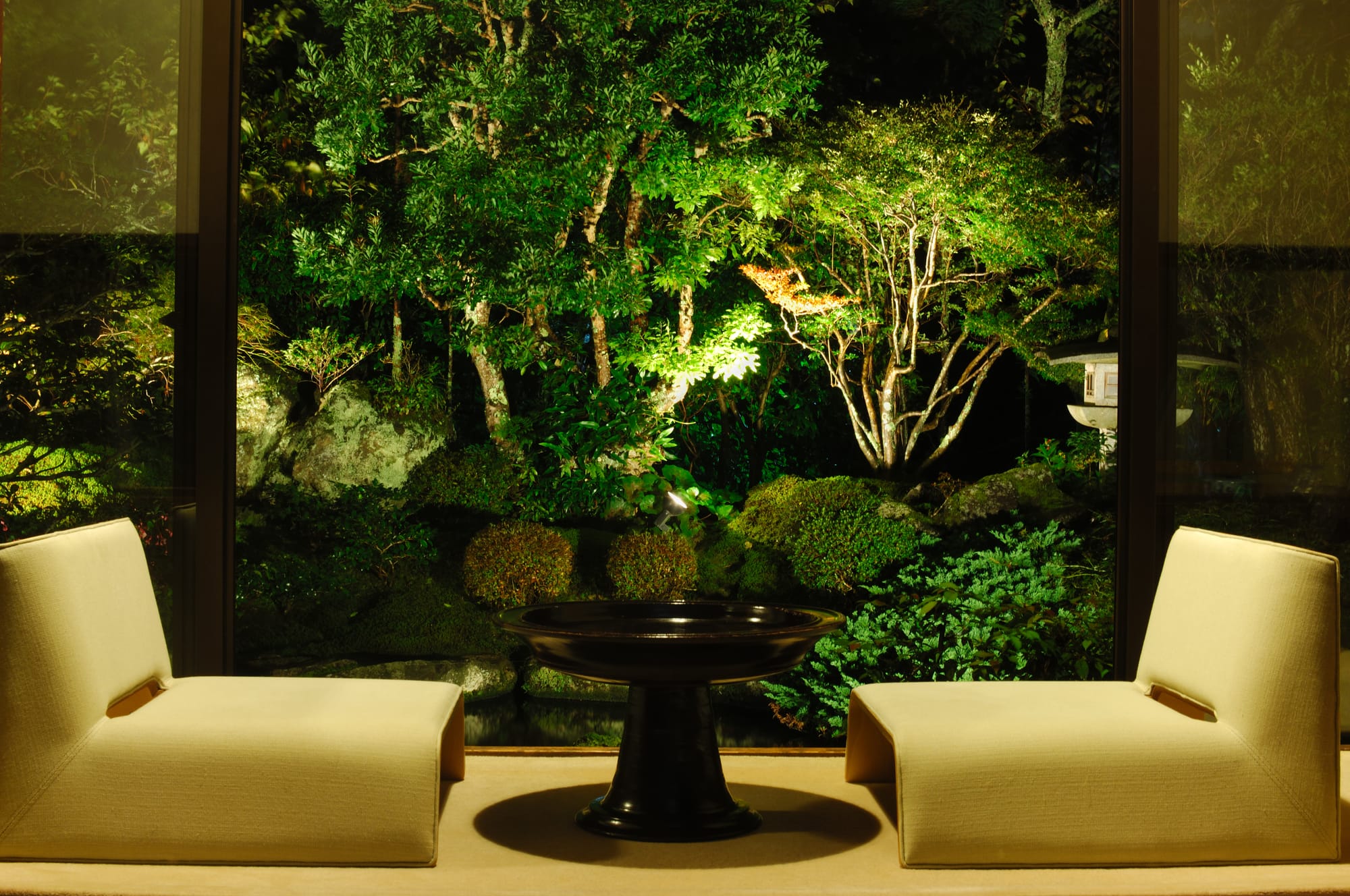Two cream lounge chairs facing each other across a low lacquer table, an illuminated Japanese garden of moss and maple beyond the glass at night