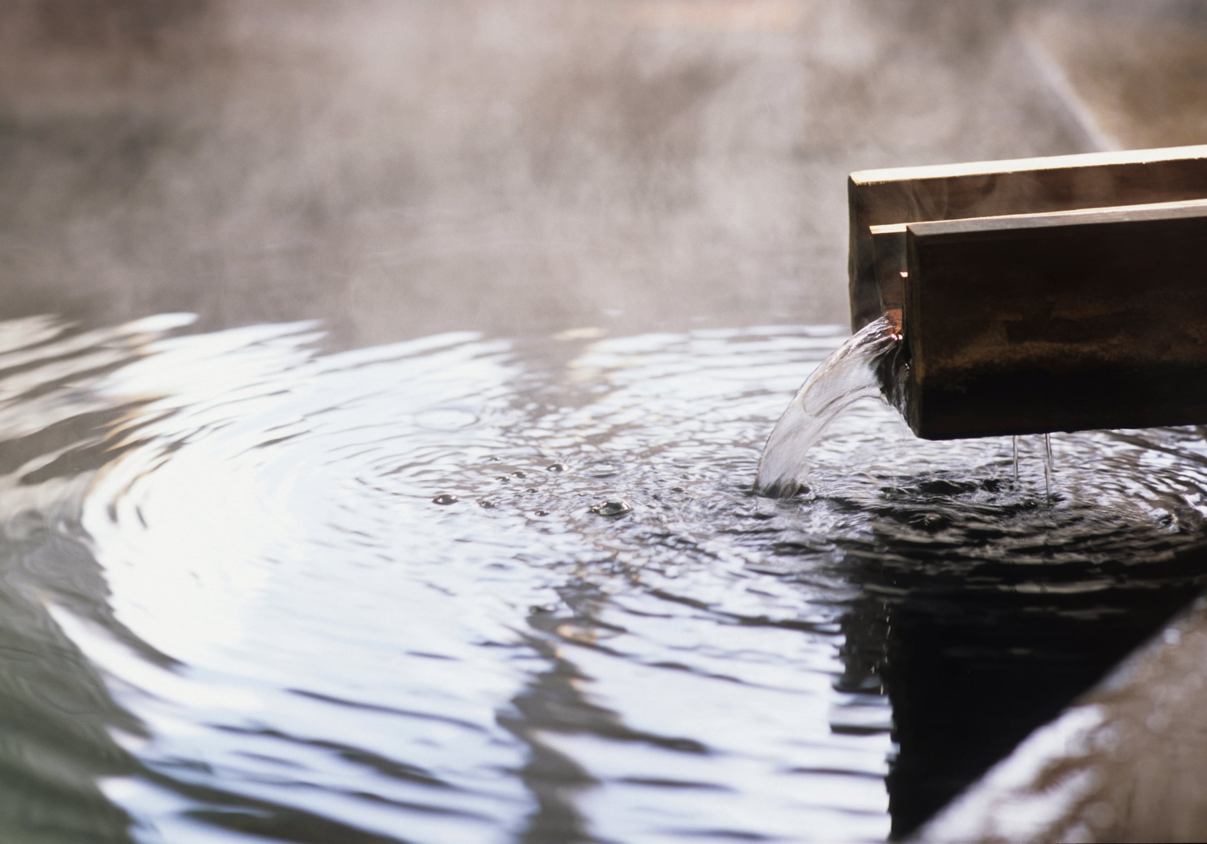 Hot spring water pouring from a wooden spout into an onsen bath, steam rising from the rippled surface