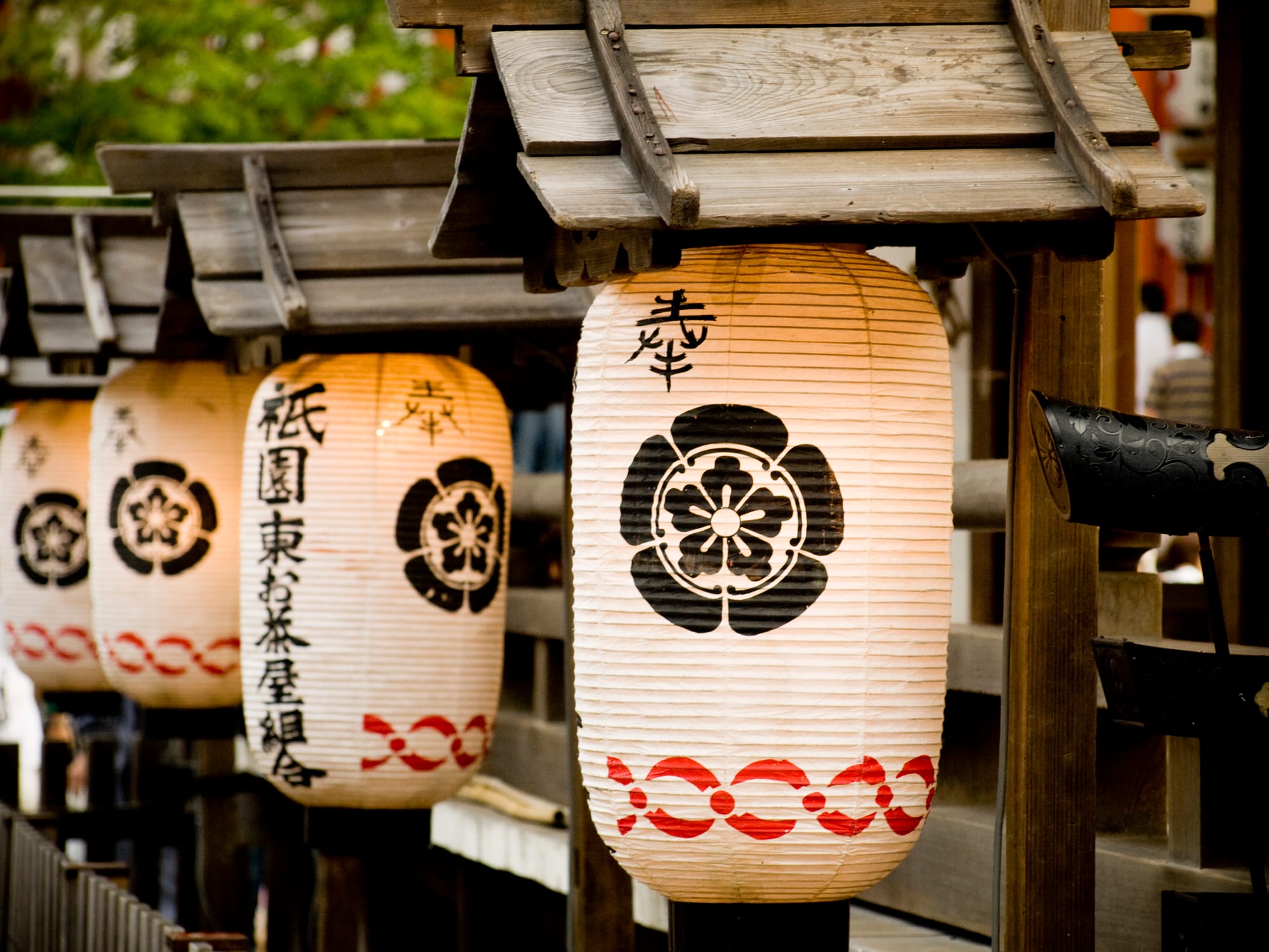 A row of lit white paper lanterns, each with a black flower crest and a red band at the foot, hanging beneath small wooden roofs on posts, one of them lettered with the name Gion Higashi