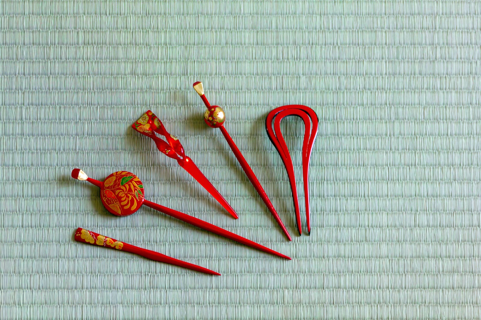 Five red lacquered pins and combs, some decorated with gold and painted flowers, laid side by side on a pale green tatami mat
