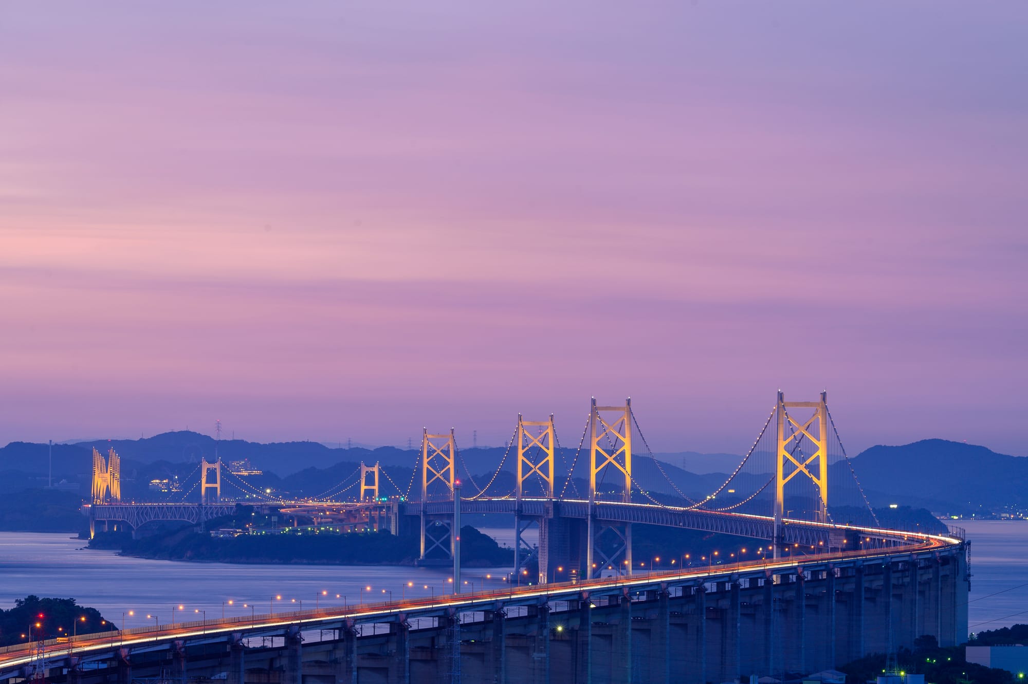 A suspension bridge crossing the Seto Inland Sea at dusk, its towers lit gold above dark water and low island silhouettes
