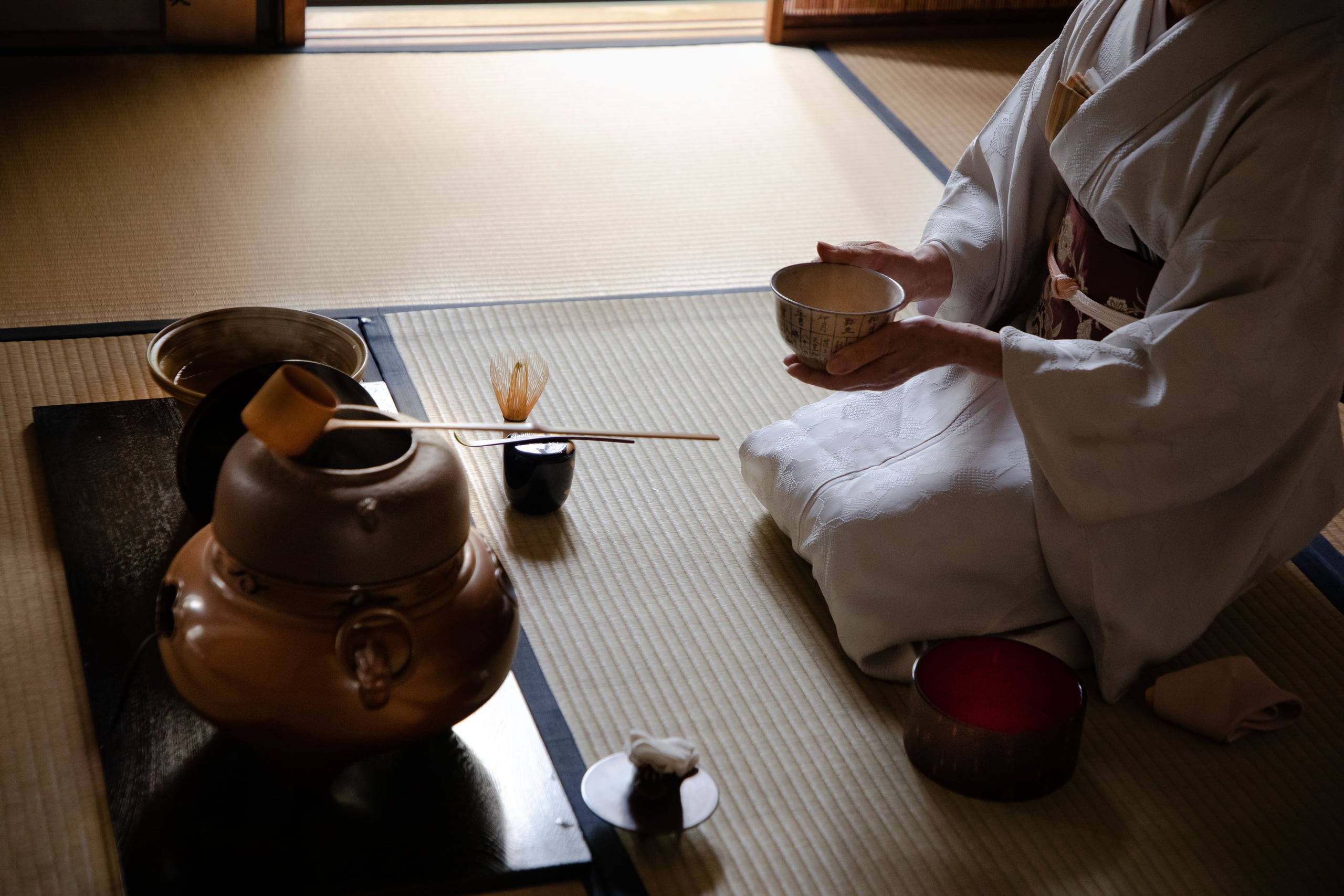 A tea host in a white kimono kneeling by a window on tatami, a tea bowl in her hands