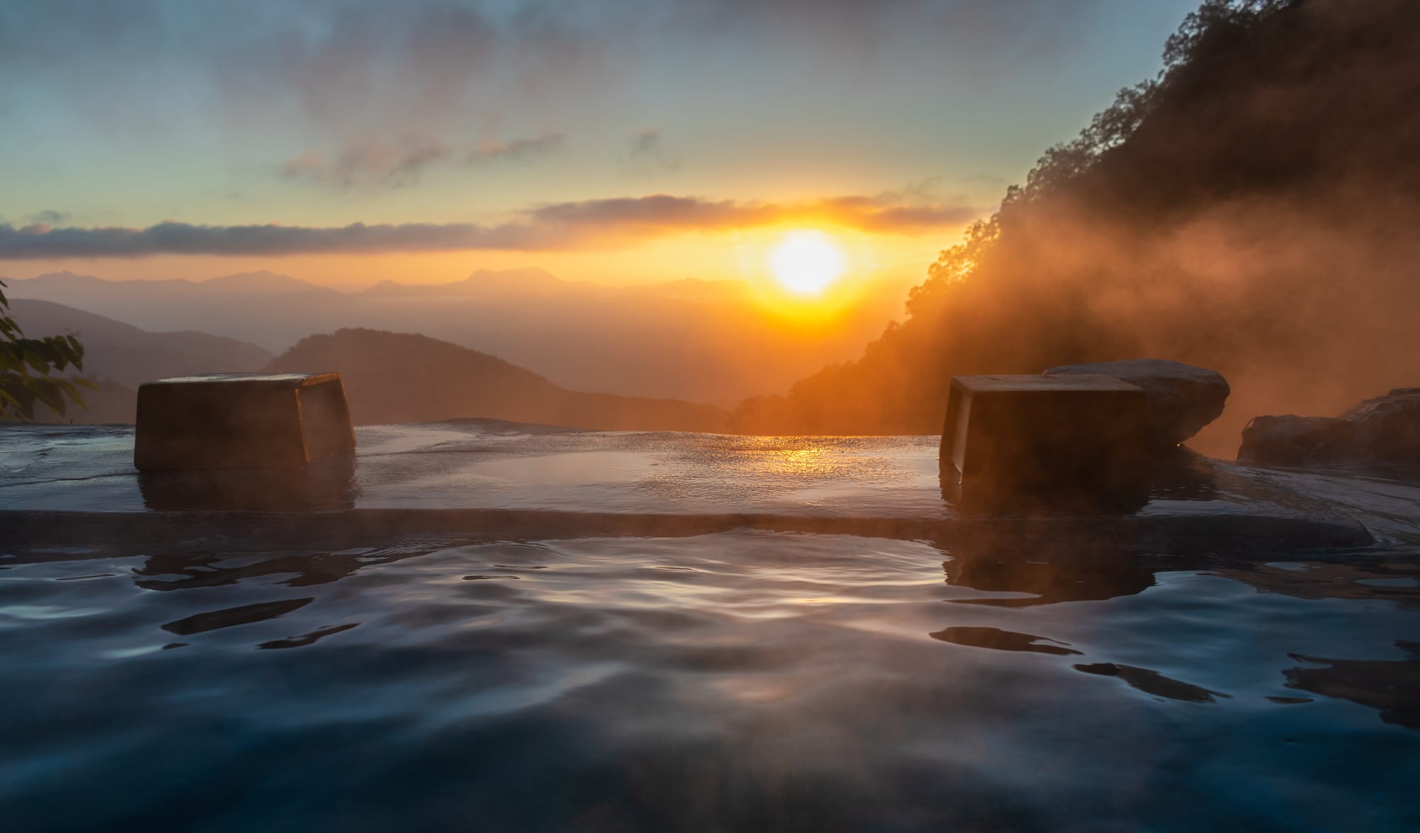 A stone open-air onsen at sunrise, steam drifting off the water and a forested slope, with ridges of mountains fading into the haze beyond
