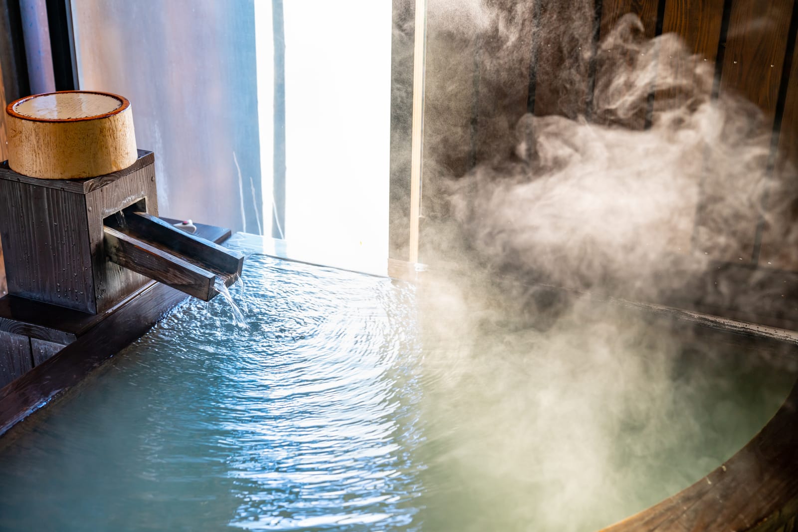 Hot spring water pouring from a wooden spout into a round wooden tub, steam rising against a bright window
