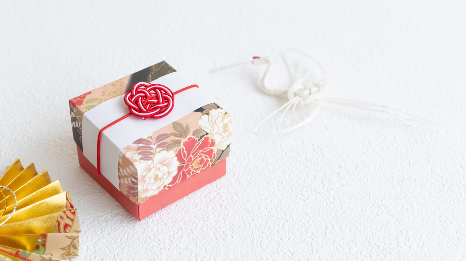 A small gift box wrapped in floral washi paper and tied with a red mizuhiki knot, beside a folded paper crane