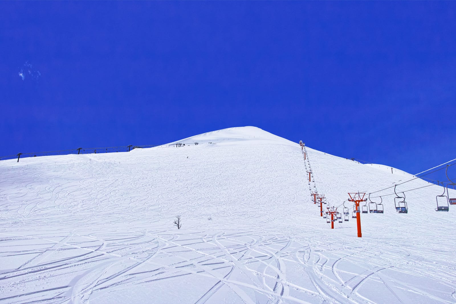 Carved ski tracks across an empty snow slope rising to a rounded summit, a red chairlift climbing beside it under a deep blue winter sky