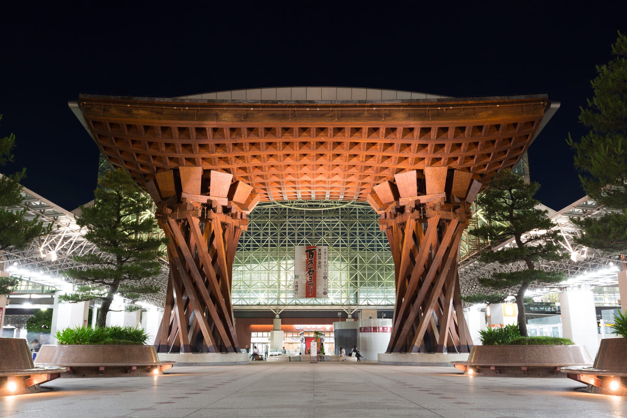 The Tsuzumi-mon gate of Kanazawa Station at night, two great twisted timber pillars holding a lattice roof, lit amber