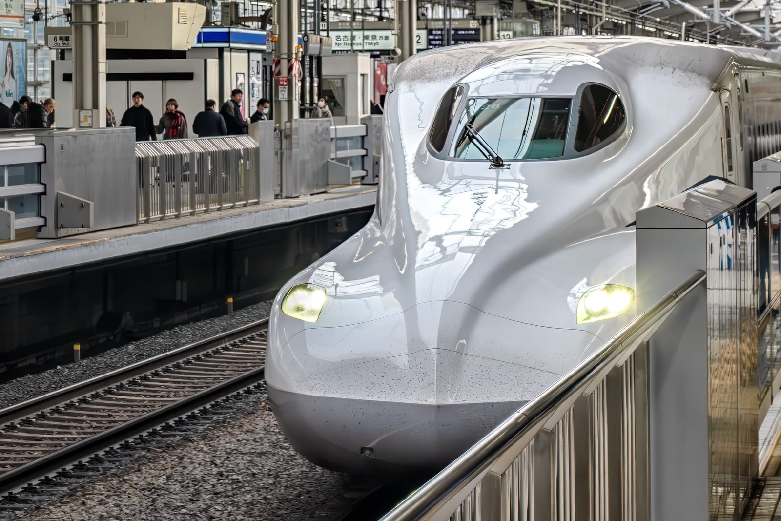 The long white nose of an N700-series shinkansen drawing into a covered station platform, headlights on