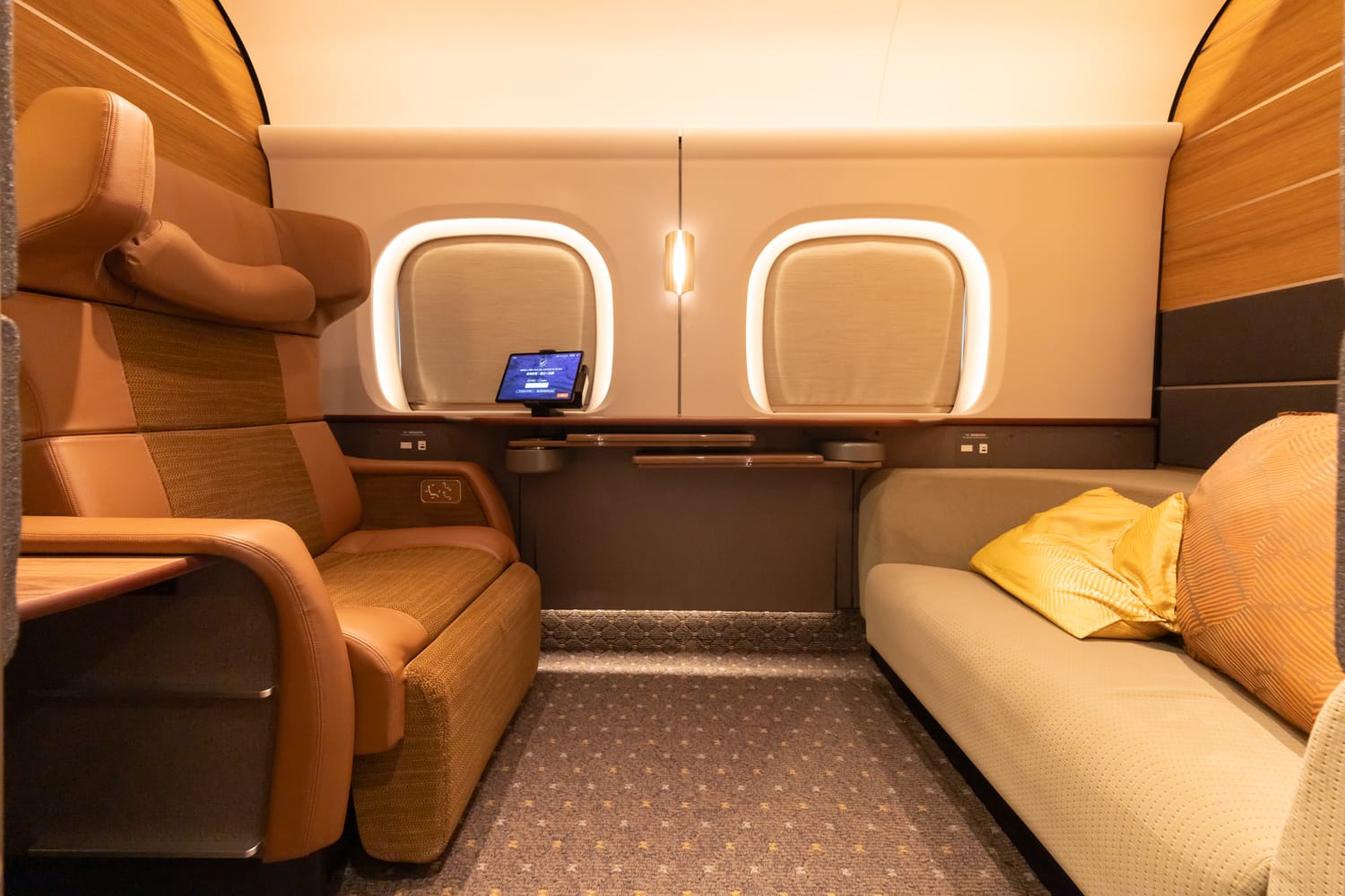 The Supreme Class cabin for two in car 7: a tan leather reclining seat on the left faces a long sofa with gold cushions, a tablet on the ledge between two softly lit rounded windows