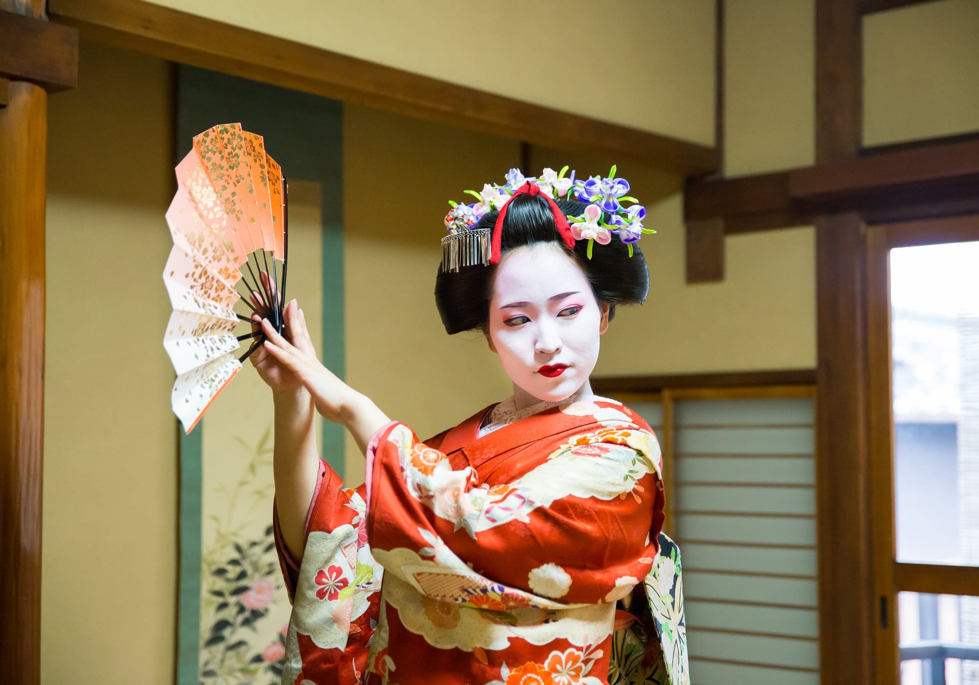 A maiko in a red kimono and flowered hair ornaments dances with an open fan in a tatami room, a painted scroll and shoji screen behind her