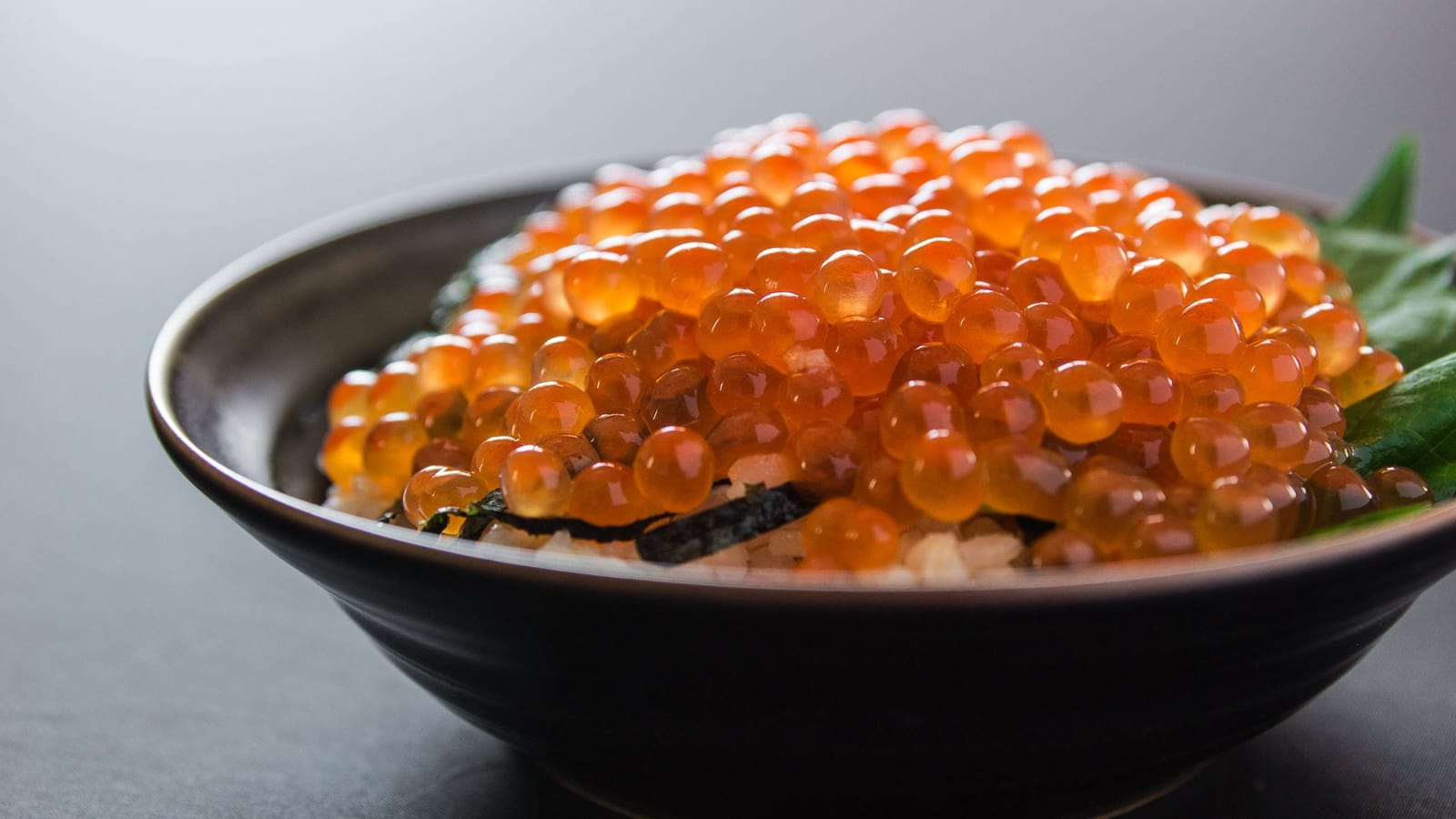 A close-up bowl of glistening orange salmon roe over rice, with nori and a shiso leaf