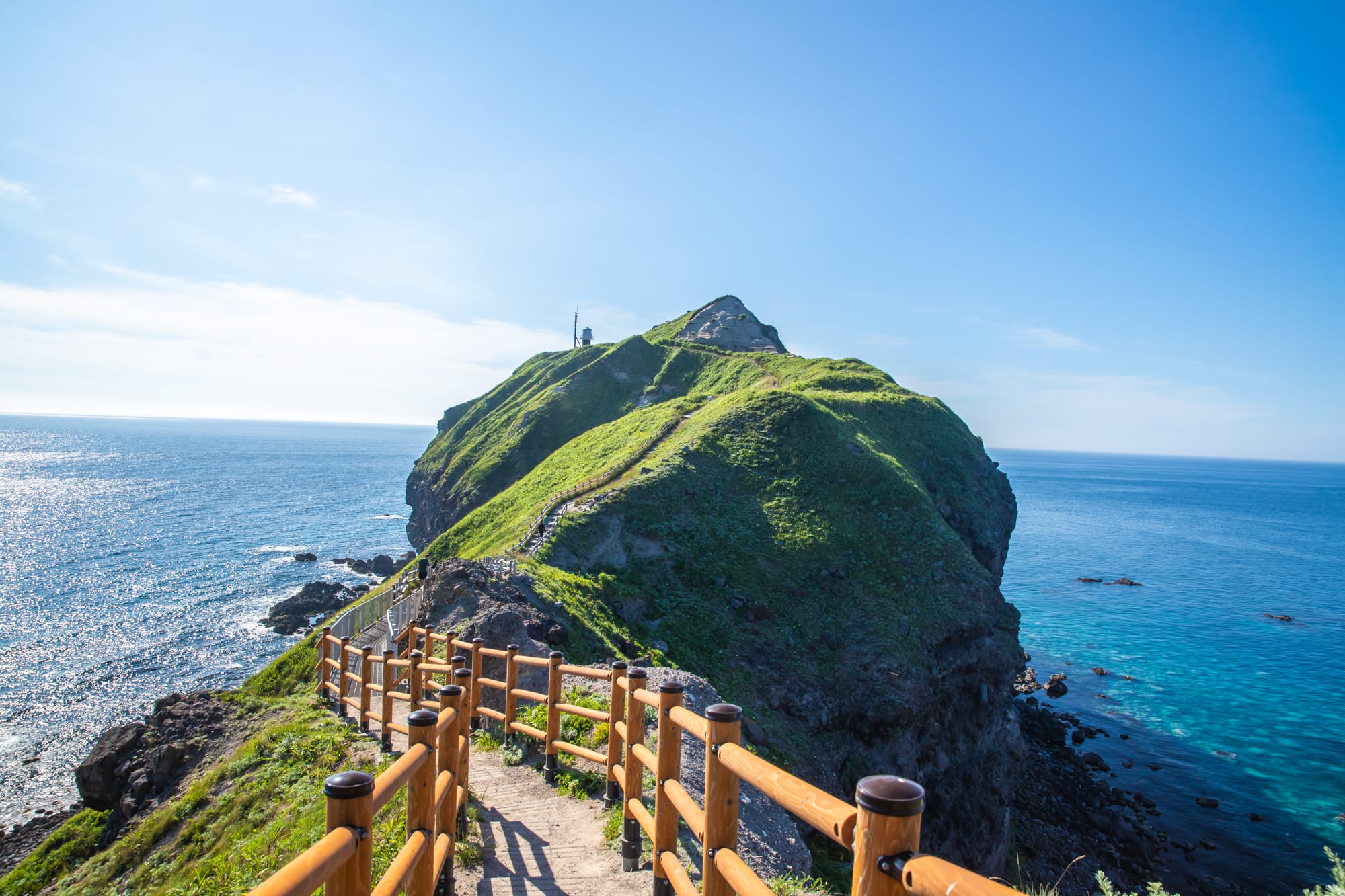 A fenced path running out along a narrow green ridge to the rocky tip of Cape Kamui, the sea clear turquoise on one side and dark blue on the other under a summer sky