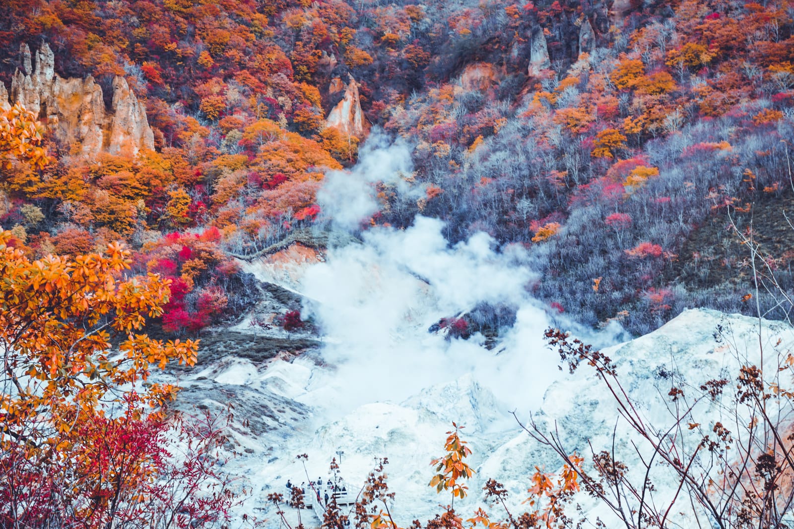 Steam rising from the pale mineral floor of Jigokudani in Noboribetsu, the slopes around it red, orange and gold with autumn leaves
