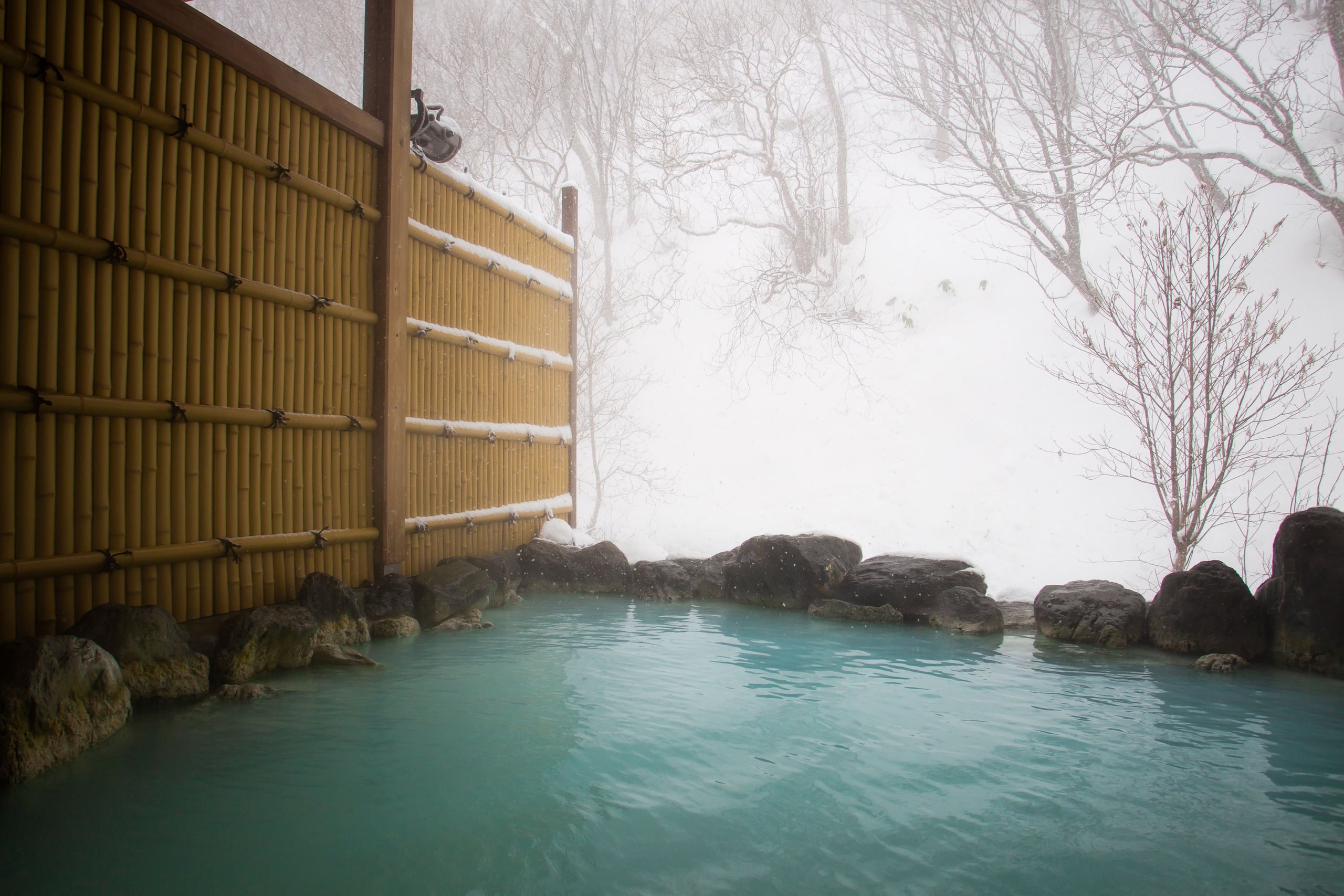 A milky turquoise rotenburo edged with dark rocks, snow falling heavily behind a bamboo fence