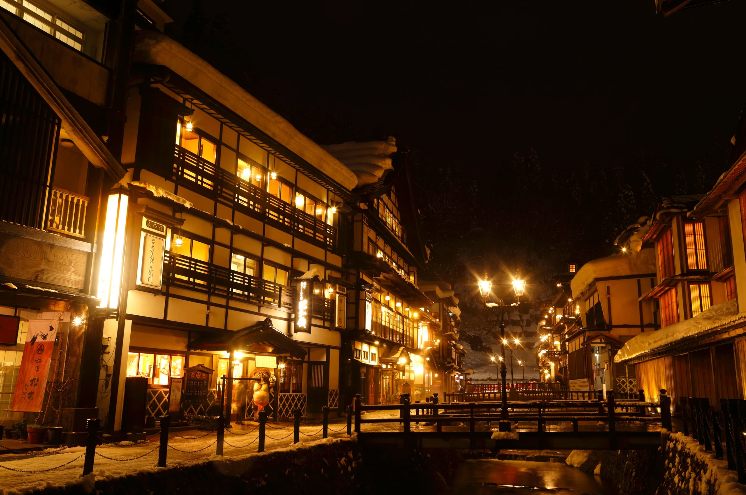 The snow-laden wooden ryokan of Ginzan Onsen facing each other across the river at night, the gas lamps lit