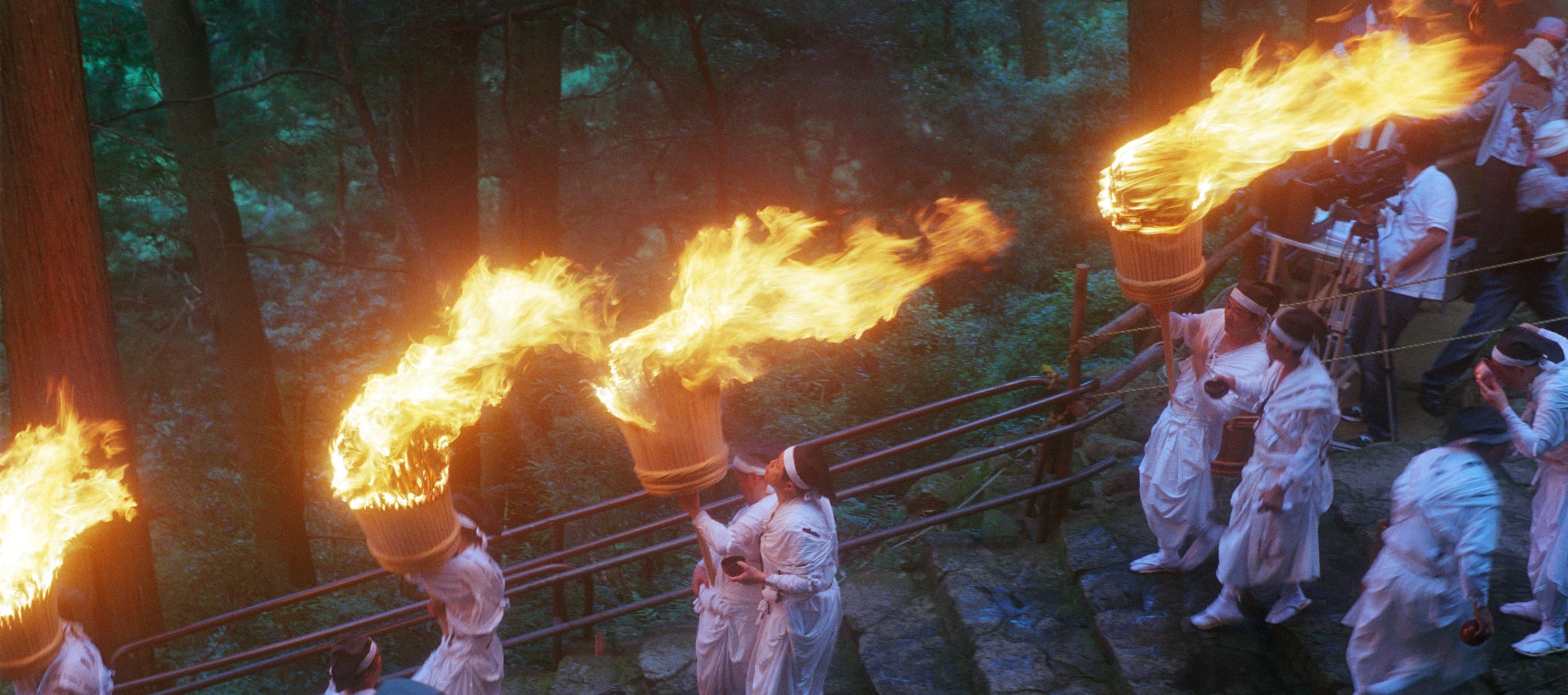 White-robed bearers in hachimaki headbands carrying giant flaming torches down stone steps at the Nachi Fire Festival, Kumano