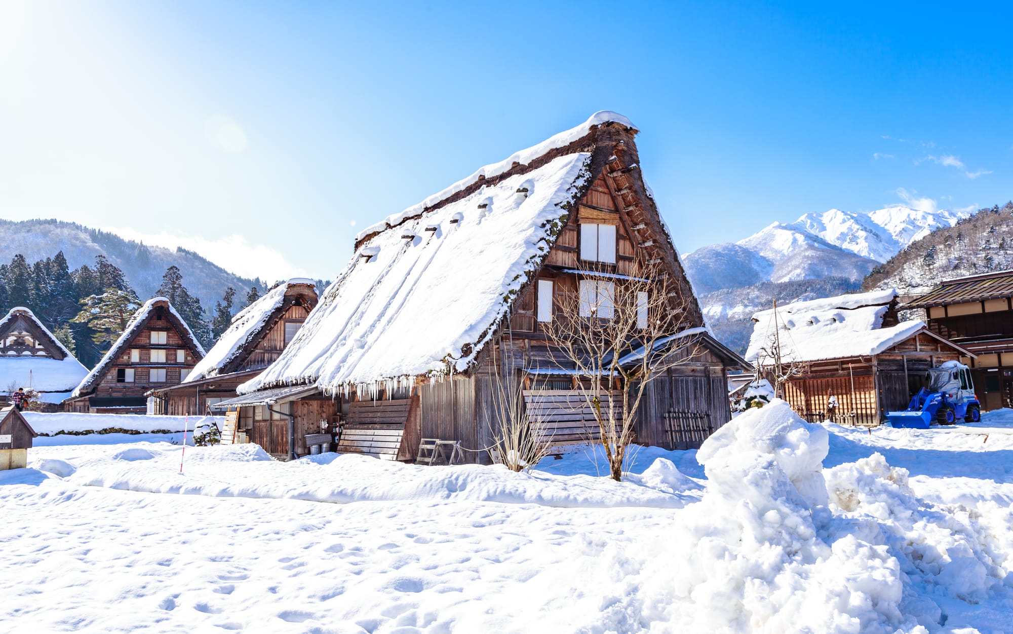 Snow-covered gassho-zukuri farmhouses with steep thatched roofs in Shirakawa-go, a snowy mountain ridge under a clear blue sky beyond