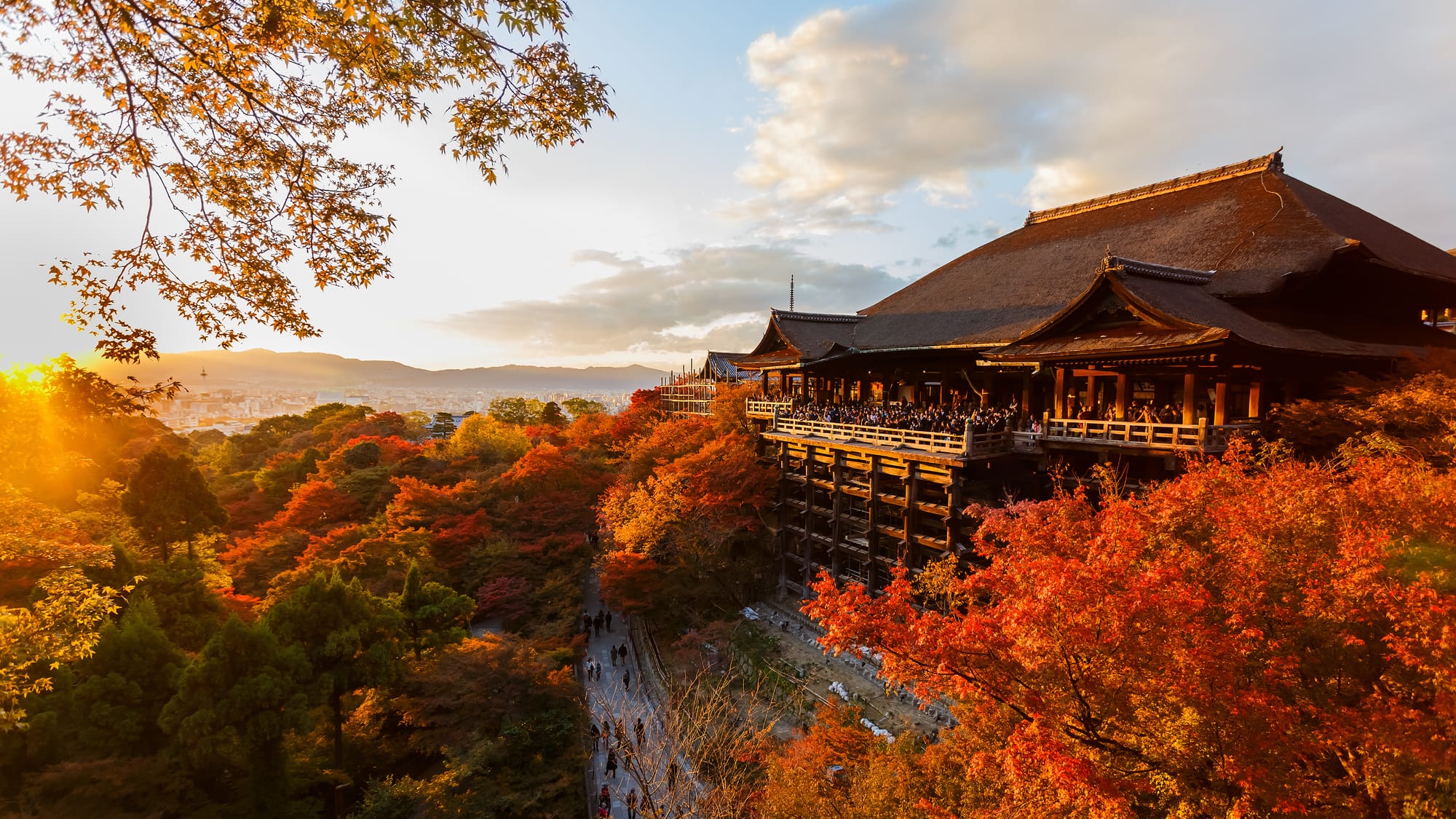 The great wooden hall and stage of Kiyomizu-dera on its hillside, red and orange maples below it and in the foreground, a low sun at the left over the city of Kyoto