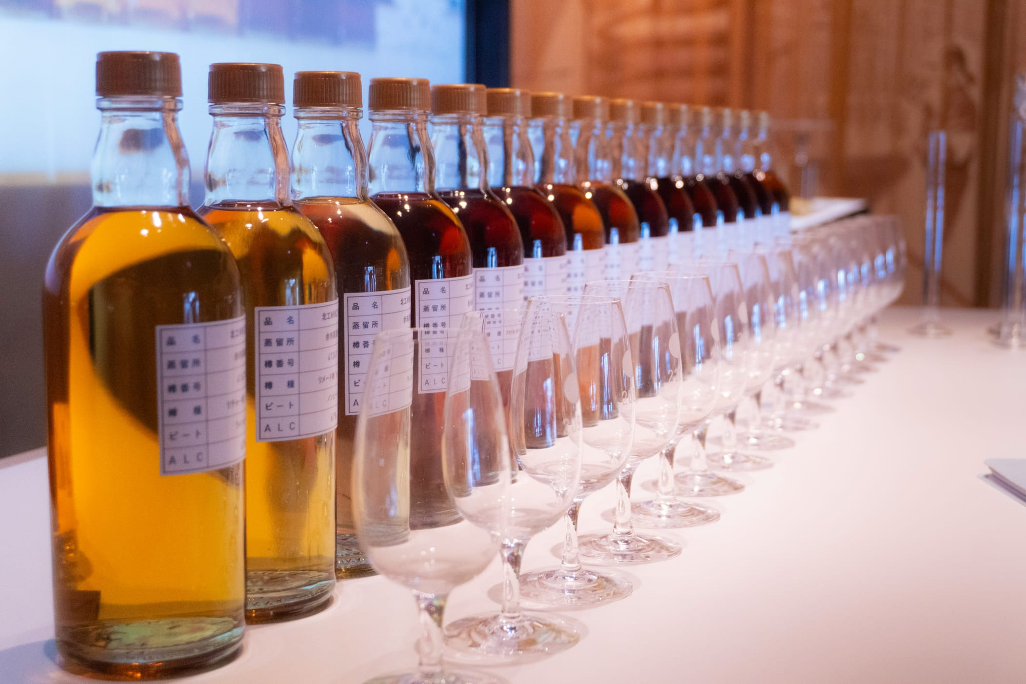 A long row of sample bottles of whisky, pale gold to deep amber, each with a nosing glass, on a white table at the Nikka distillery in Yoichi