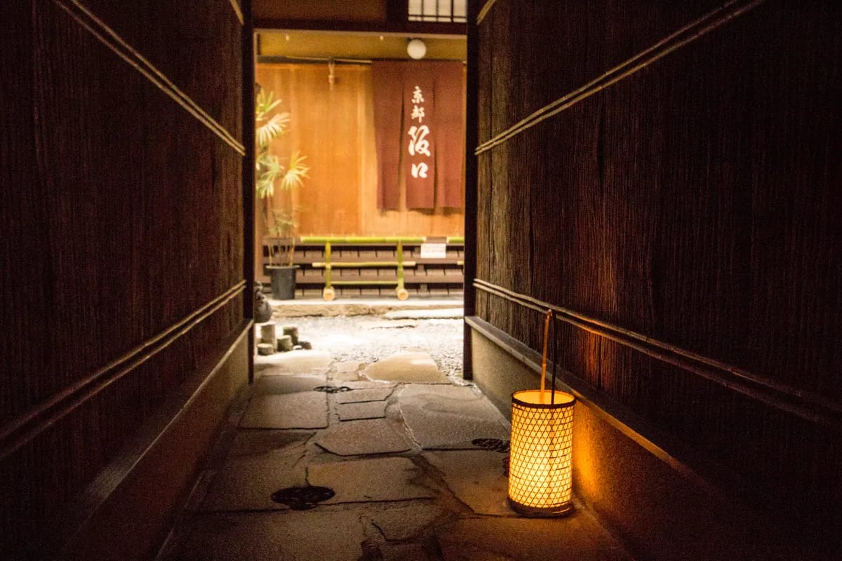 A narrow wooden teahouse entrance in Kyoto lit by a single paper lantern, a noren curtain visible beyond