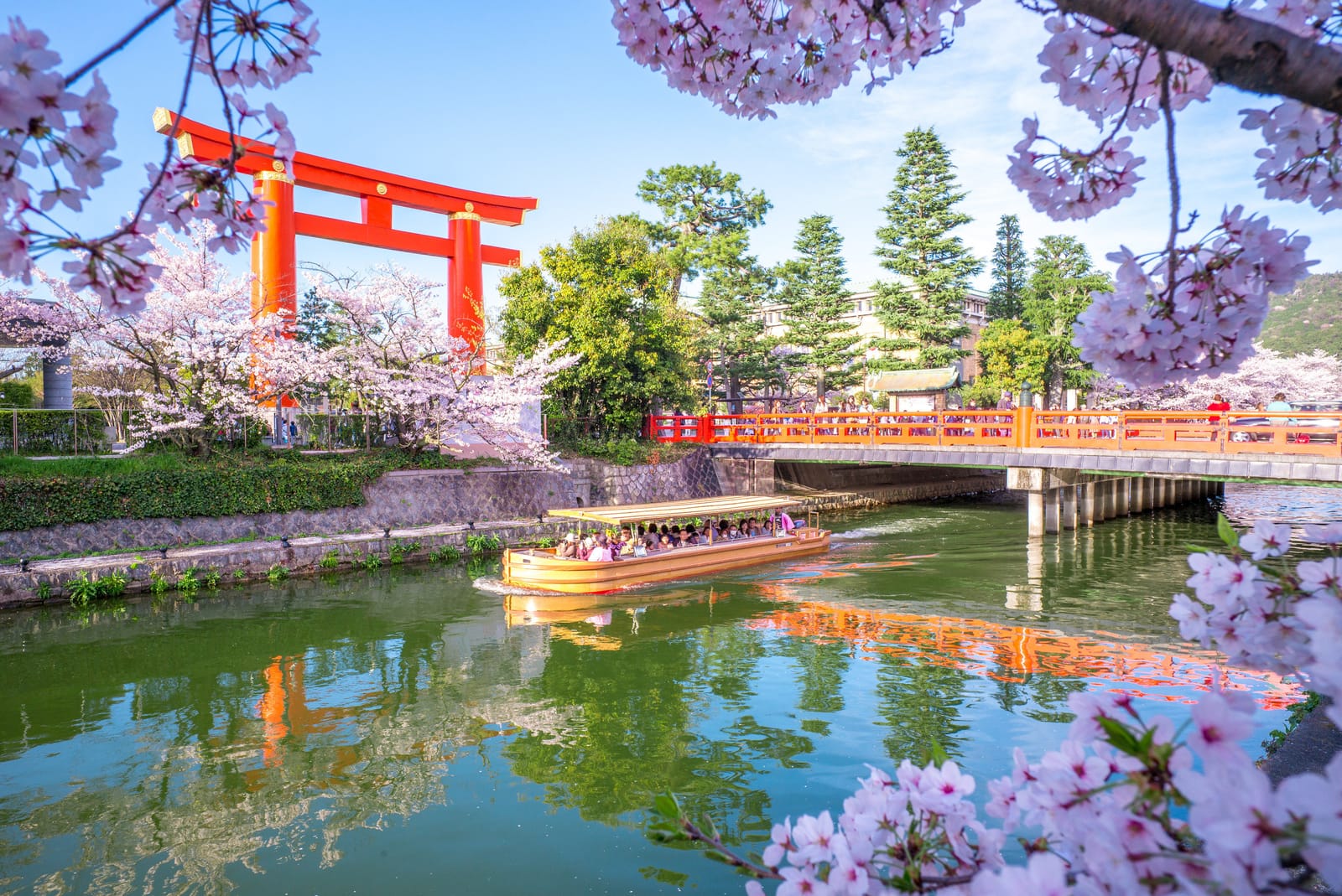 A traditional flat-bottomed canal boat carries passengers beneath the great vermilion torii of Heian Jingu on the Okazaki Canal, framed by cherry blossom branches