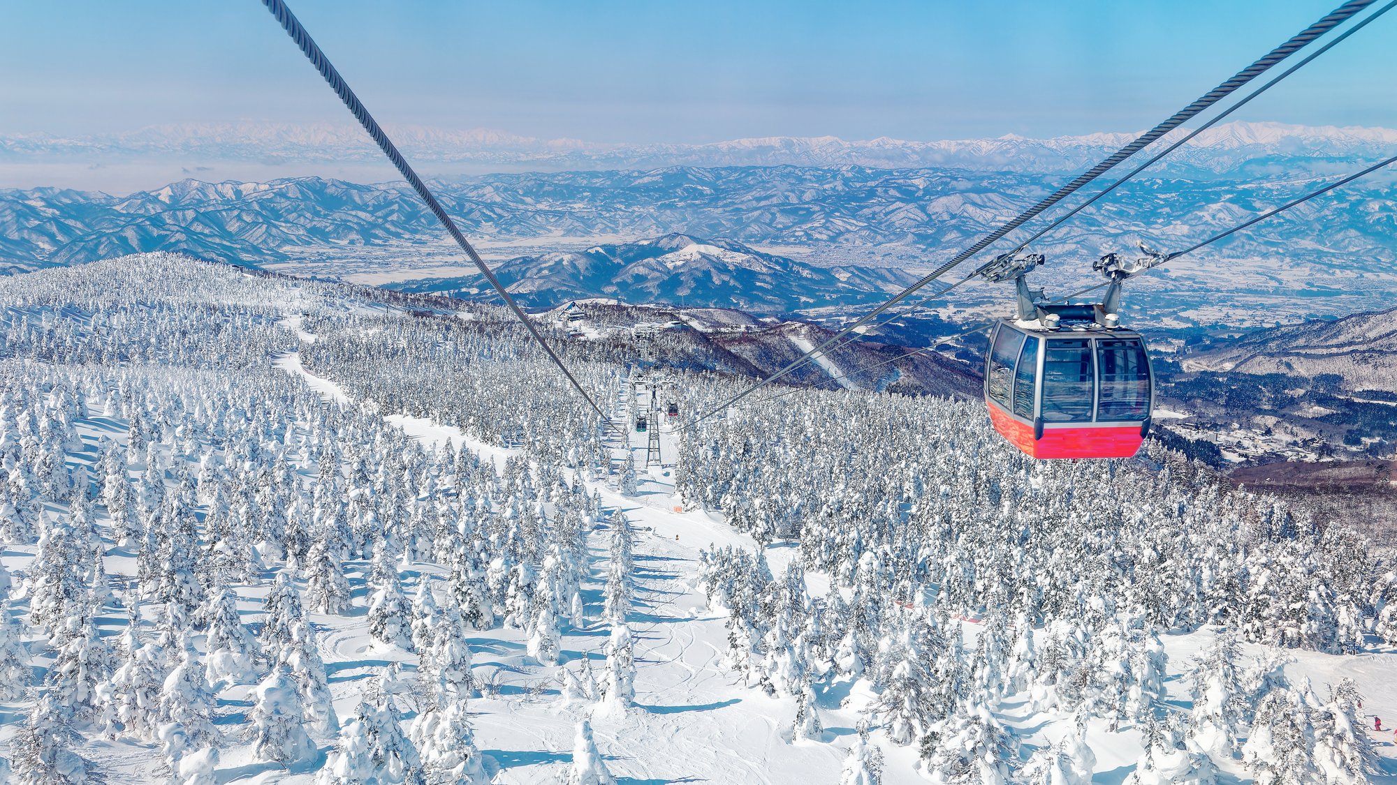 A red gondola cabin crossing above a ski run cut through snow-laden conifers, ranges of snow-covered mountains beyond
