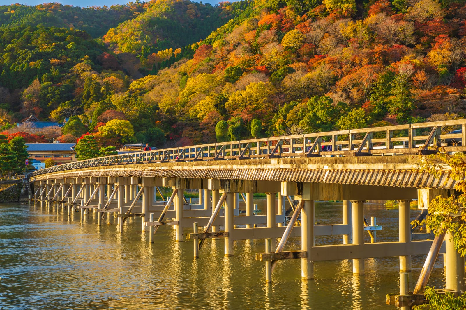 The long wooden Togetsukyo bridge crossing the Katsura River at Arashiyama, its piers reflected, autumn hillsides of red and gold behind