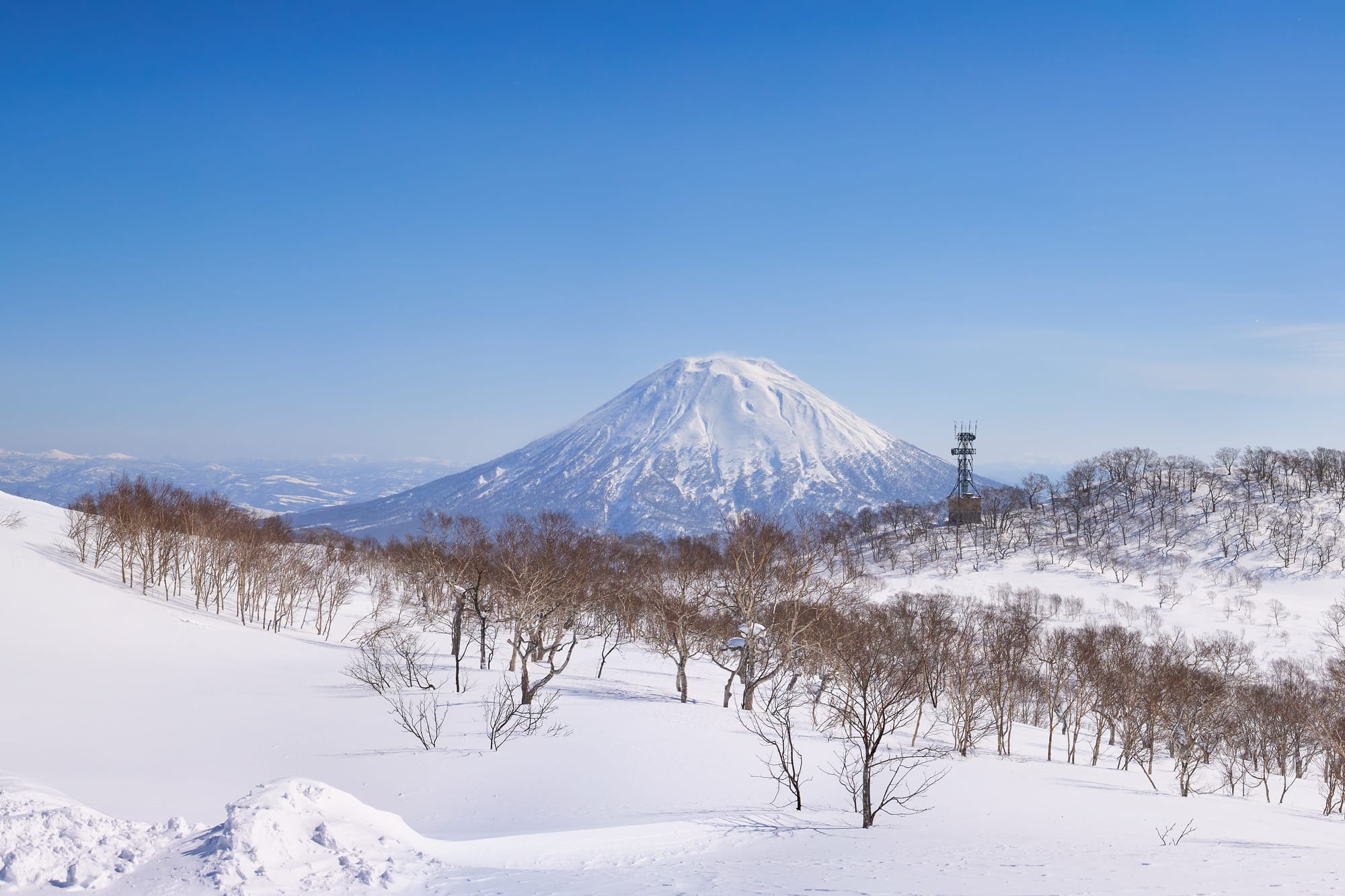 Mount Yōtei, its cone covered in snow, rising beyond bare trees and a ski lift above the slopes of Niseko under a clear blue sky