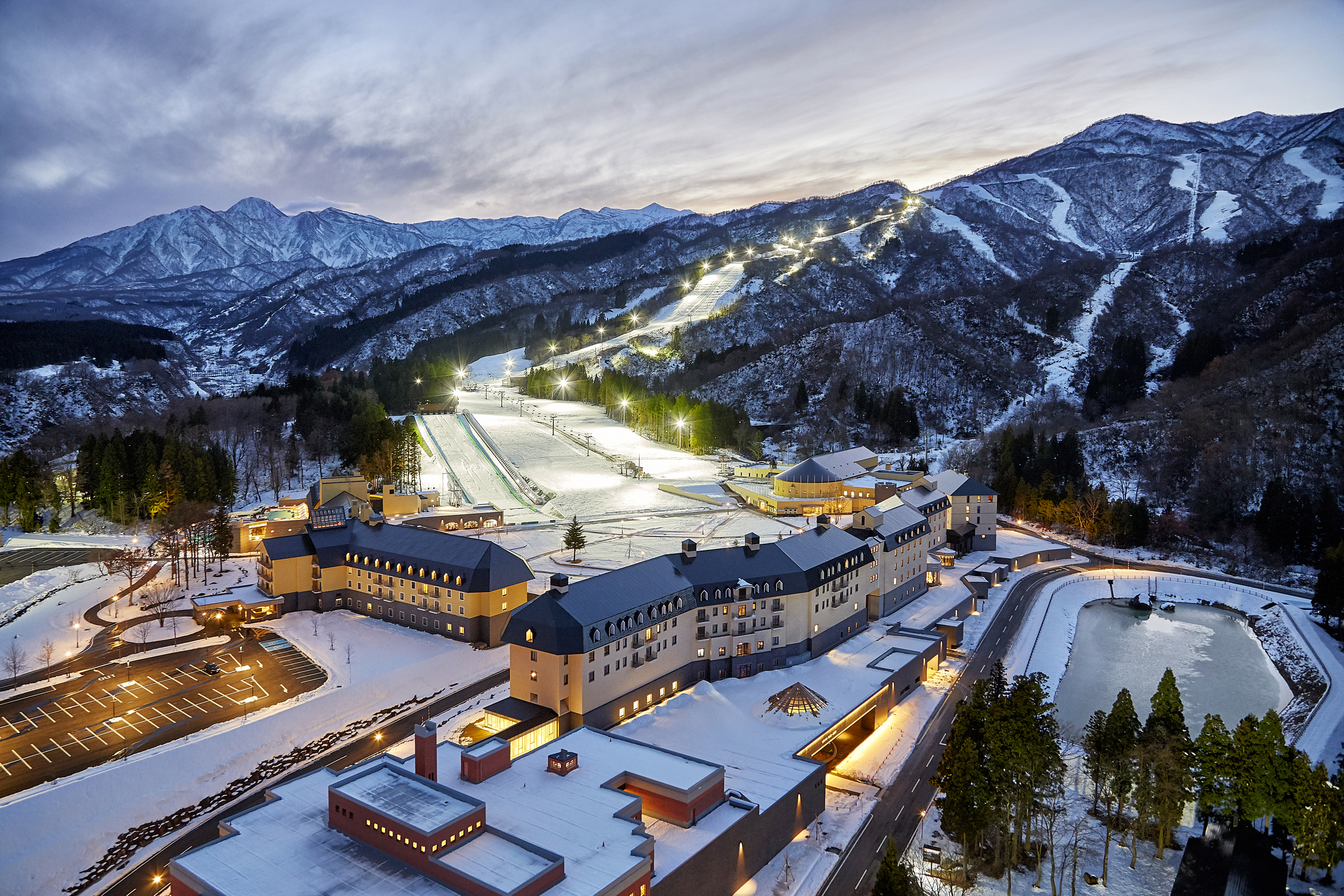 Lotte Arai Resort at dusk in winter, its two snow-covered buildings below floodlit ski slopes and a range of snowy peaks