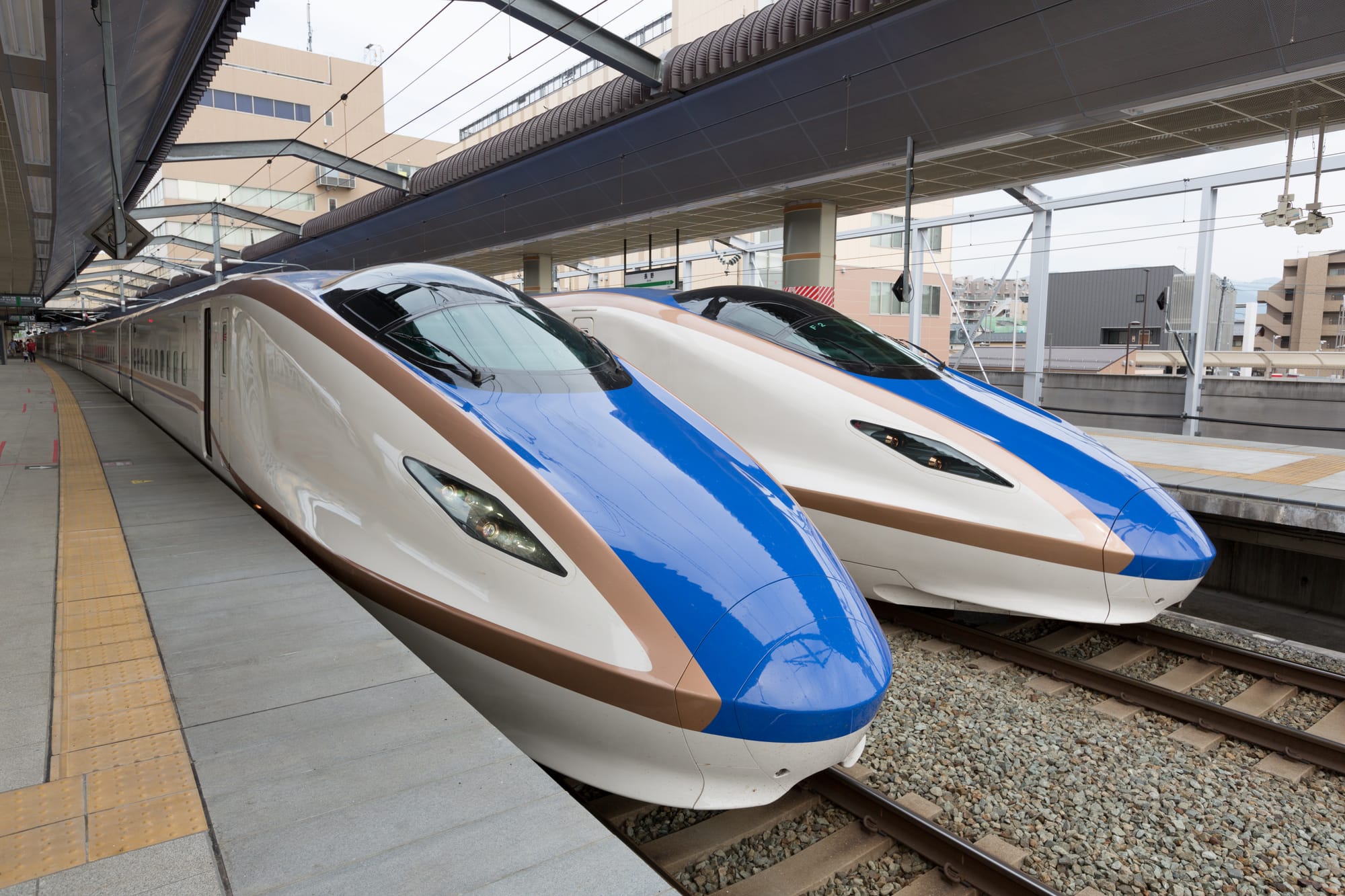 Two blue and white shinkansen bullet trains with gold trim standing side by side at a station platform beneath an overhead canopy