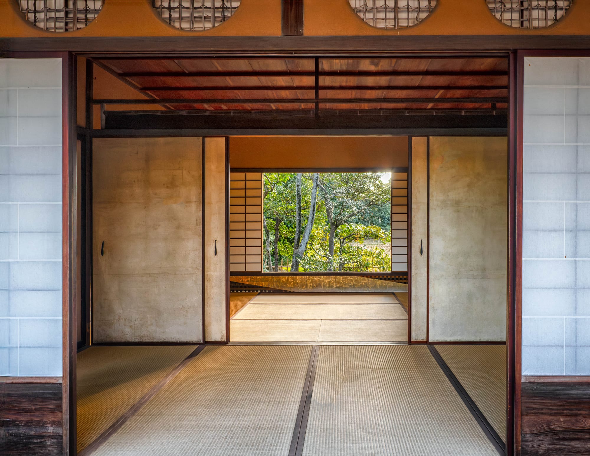A run of tatami rooms in an old Japanese house seen through open sliding doors, ending in a window full of green garden