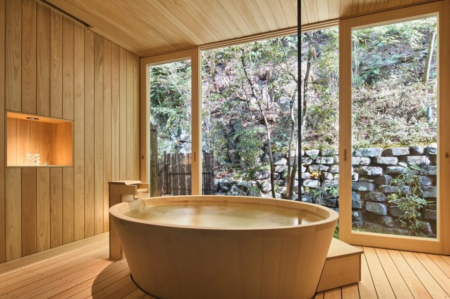 A round hinoki cypress bath filling with hot water in a private Kyoto ryokan room, a stone-walled garden visible through the glass beyond