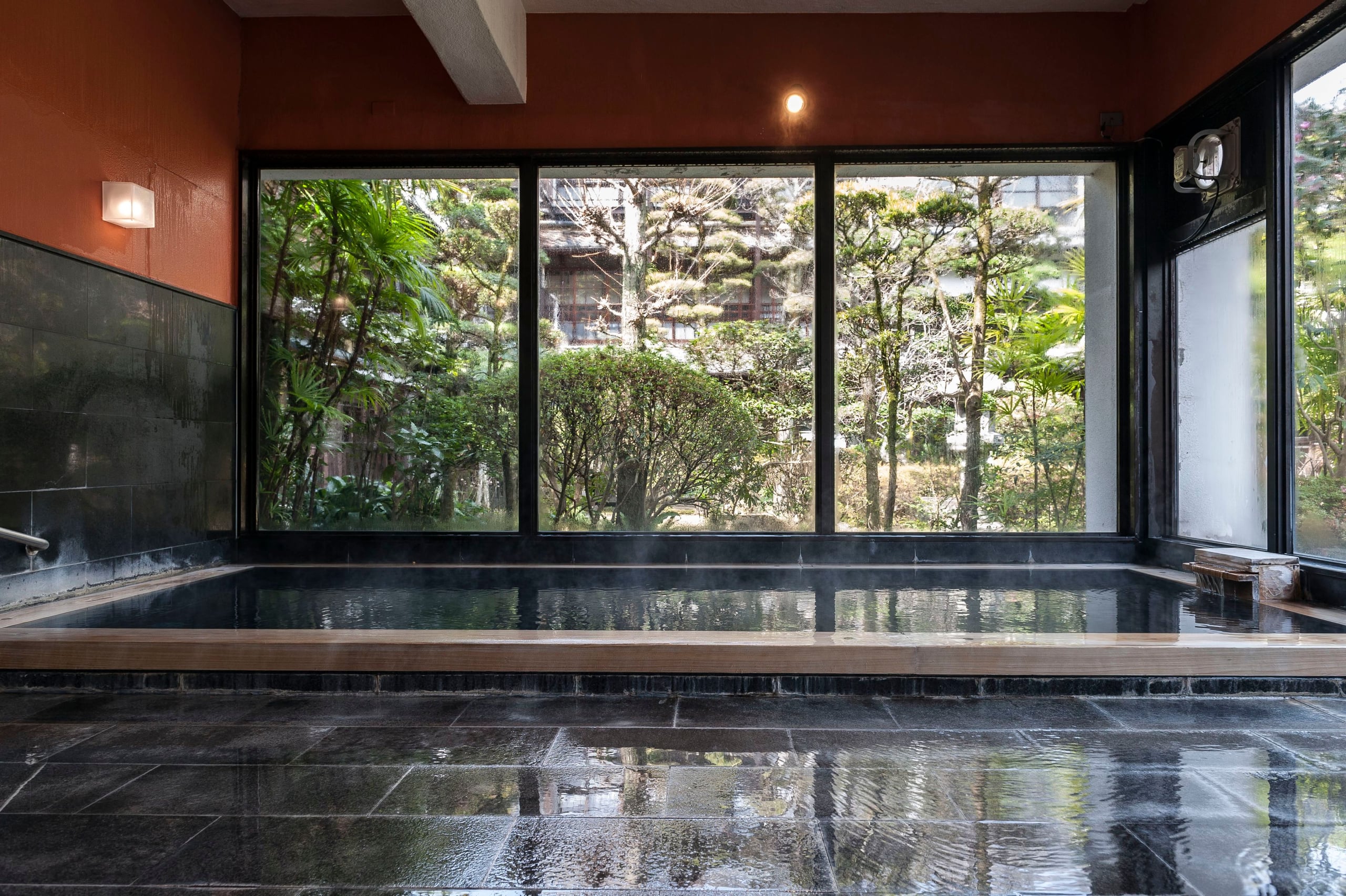 A stone-tiled indoor onsen bath steaming before a glass wall onto forest