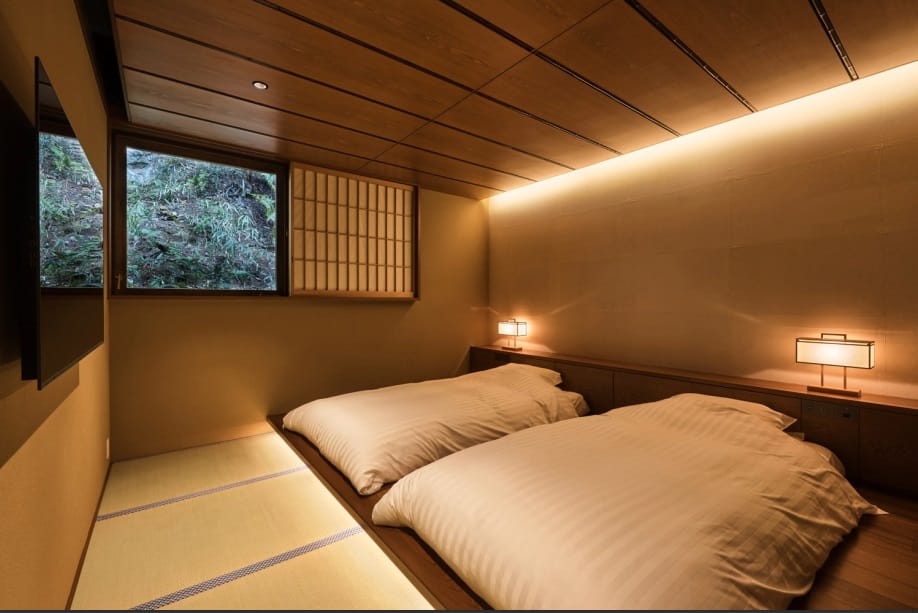 A tatami bedroom with two futon laid out beneath a shoji-framed window looking onto a wooded hillside
