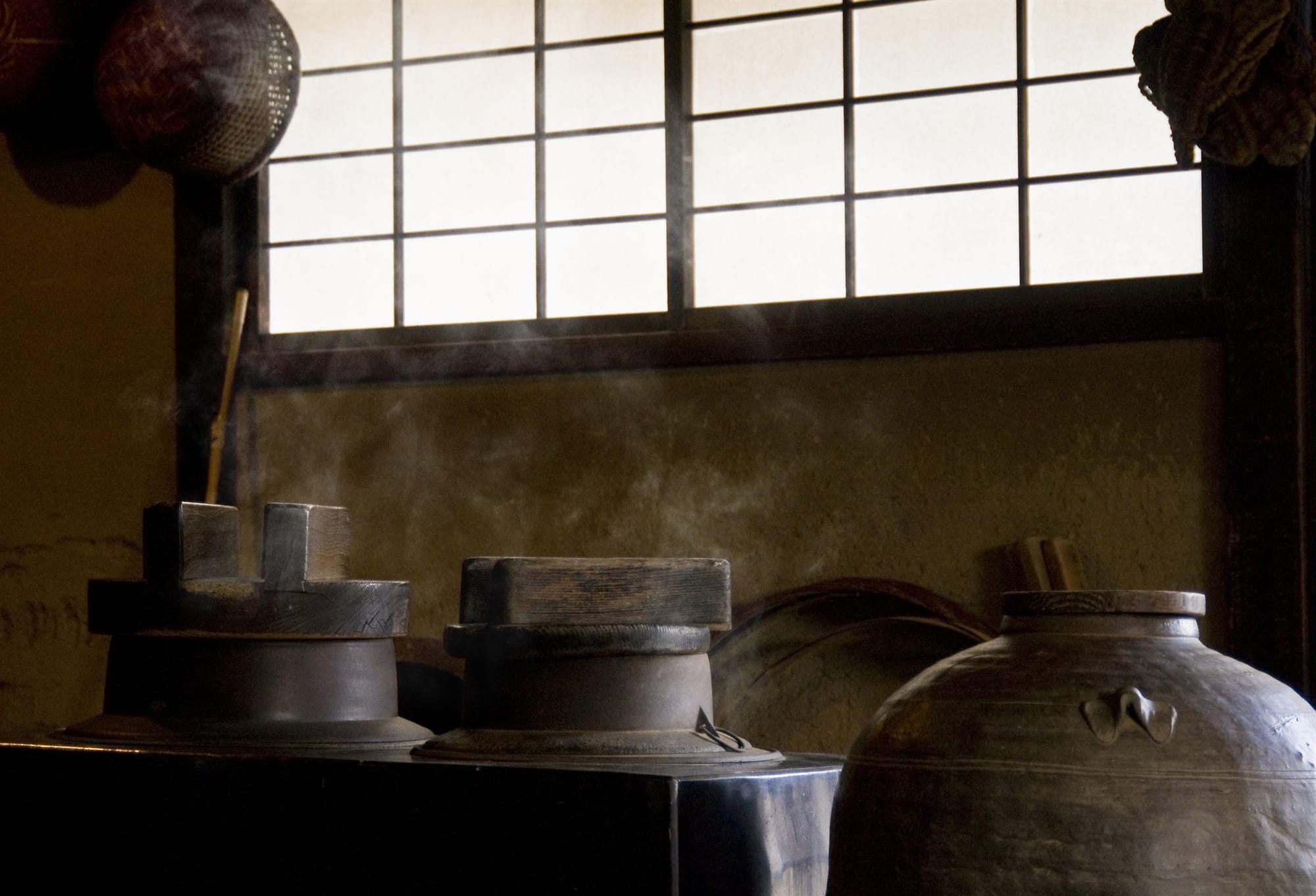 Traditional wooden sake fermentation vessels beneath a paper shoji window in a dim, steam-filled brewery interior