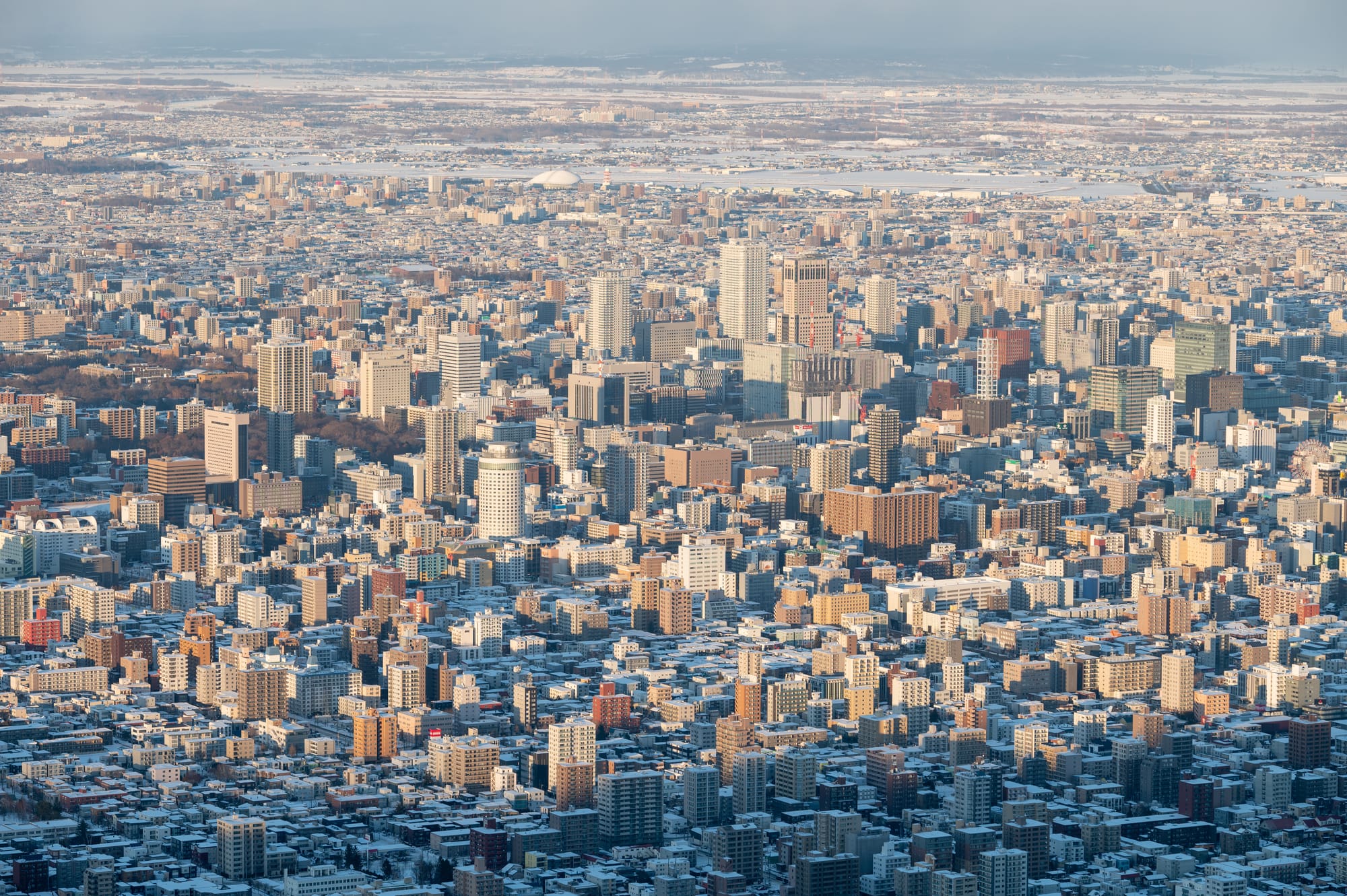 Sapporo under snow seen from high above, the towers of the city centre catching low winter light and the white plain stretching to the horizon beyond