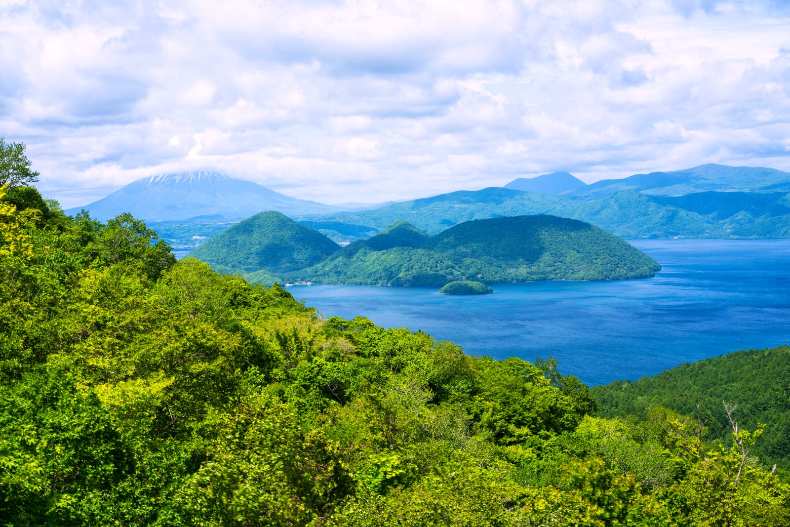 Wooded green ridges falling to the still blue water of Lake Tōya with volcanic peaks in the distance