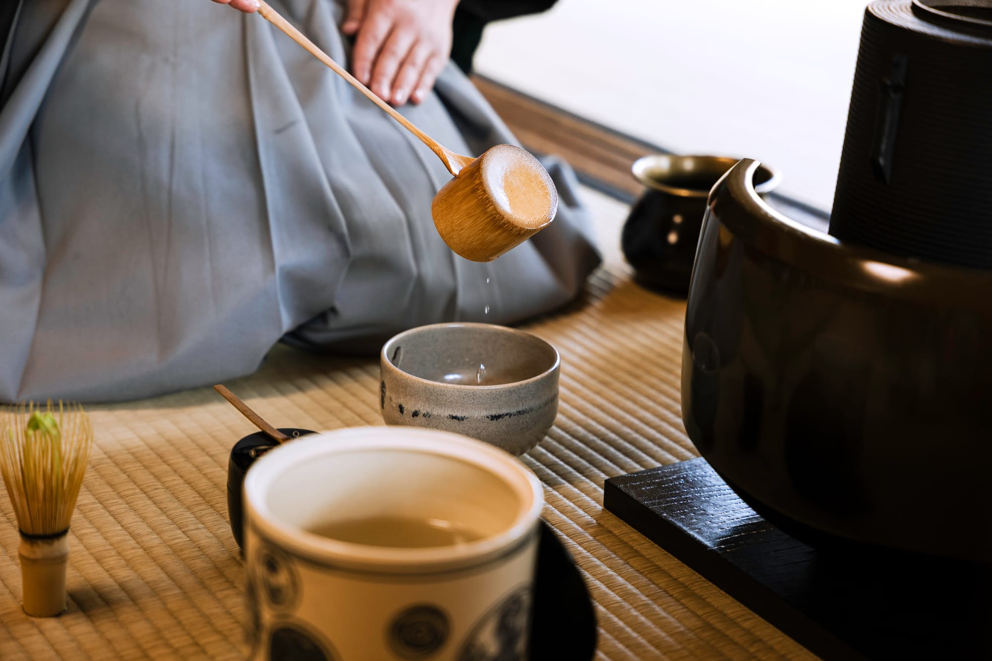 A host in a pale grey-blue kimono kneels on tatami and lifts a bamboo ladle of hot water from a black iron kettle; in the foreground a tea bowl holds green tea beside a bamboo whisk